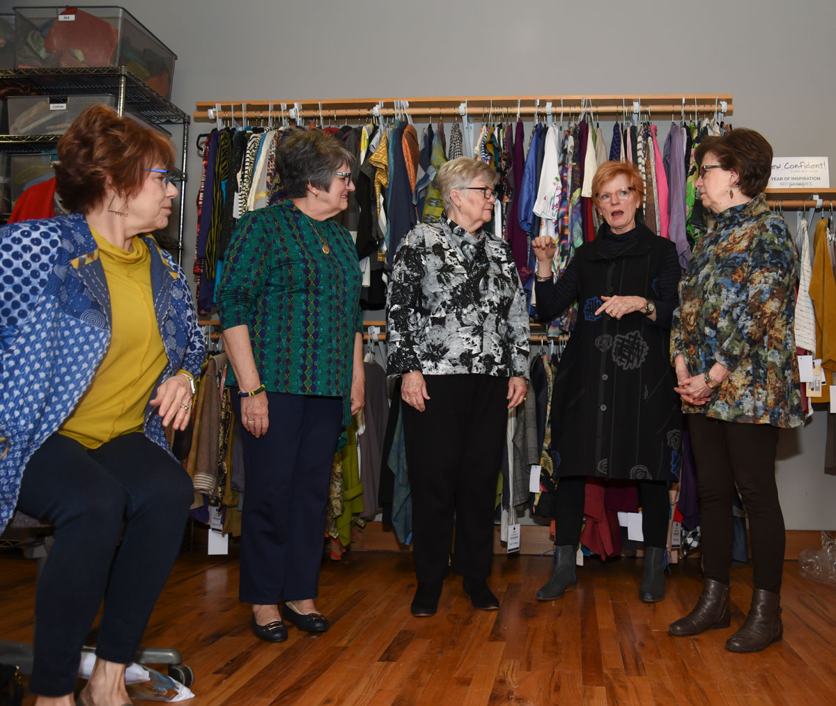 Five women stand in a clothing store, discussing fashion, with racks of clothes behind them.