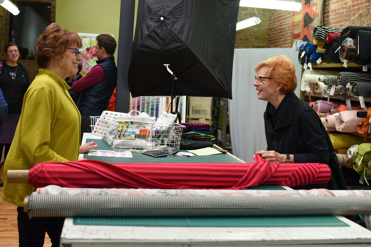 Two women with red hair and glasses talking at a market stall with fabric rolls and accessories, indoors.