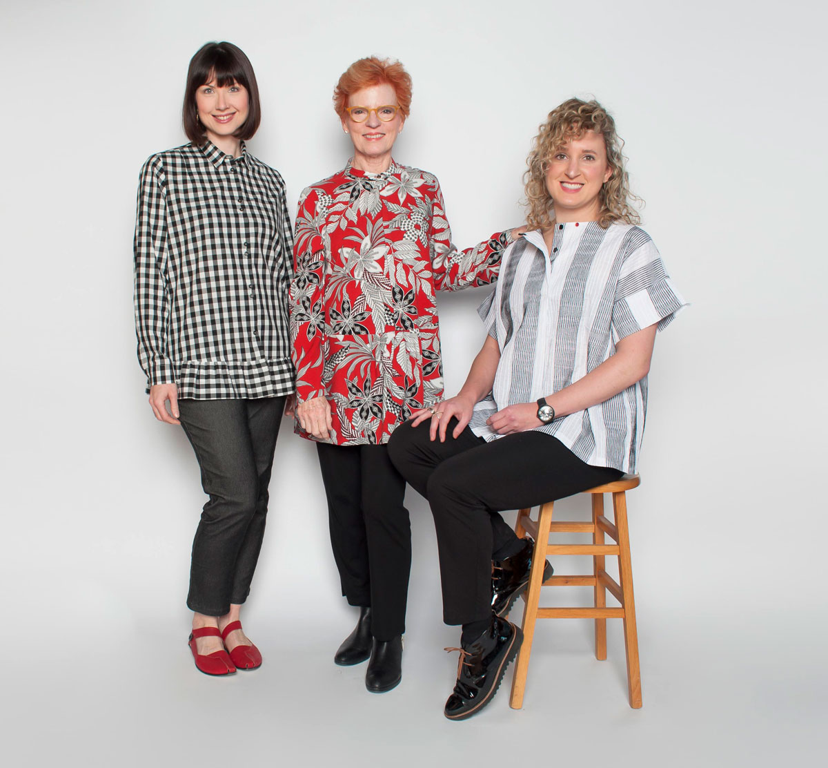 Three women standing and sitting against a plain background, one sitting on a wooden stool, all smiling.