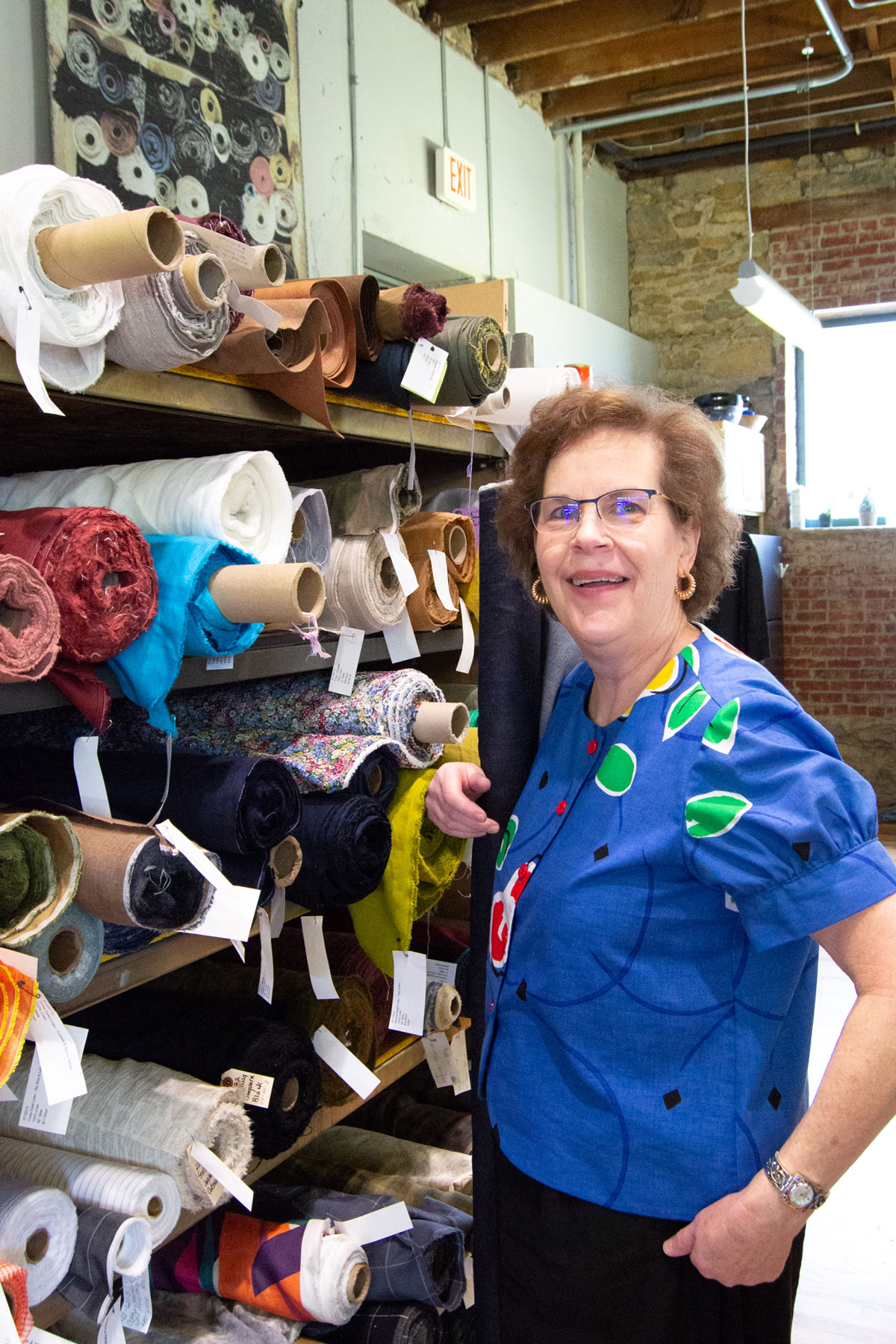Woman with glasses and curly hair smiling in fabric store, surrounded by shelves of colorful fabric rolls.