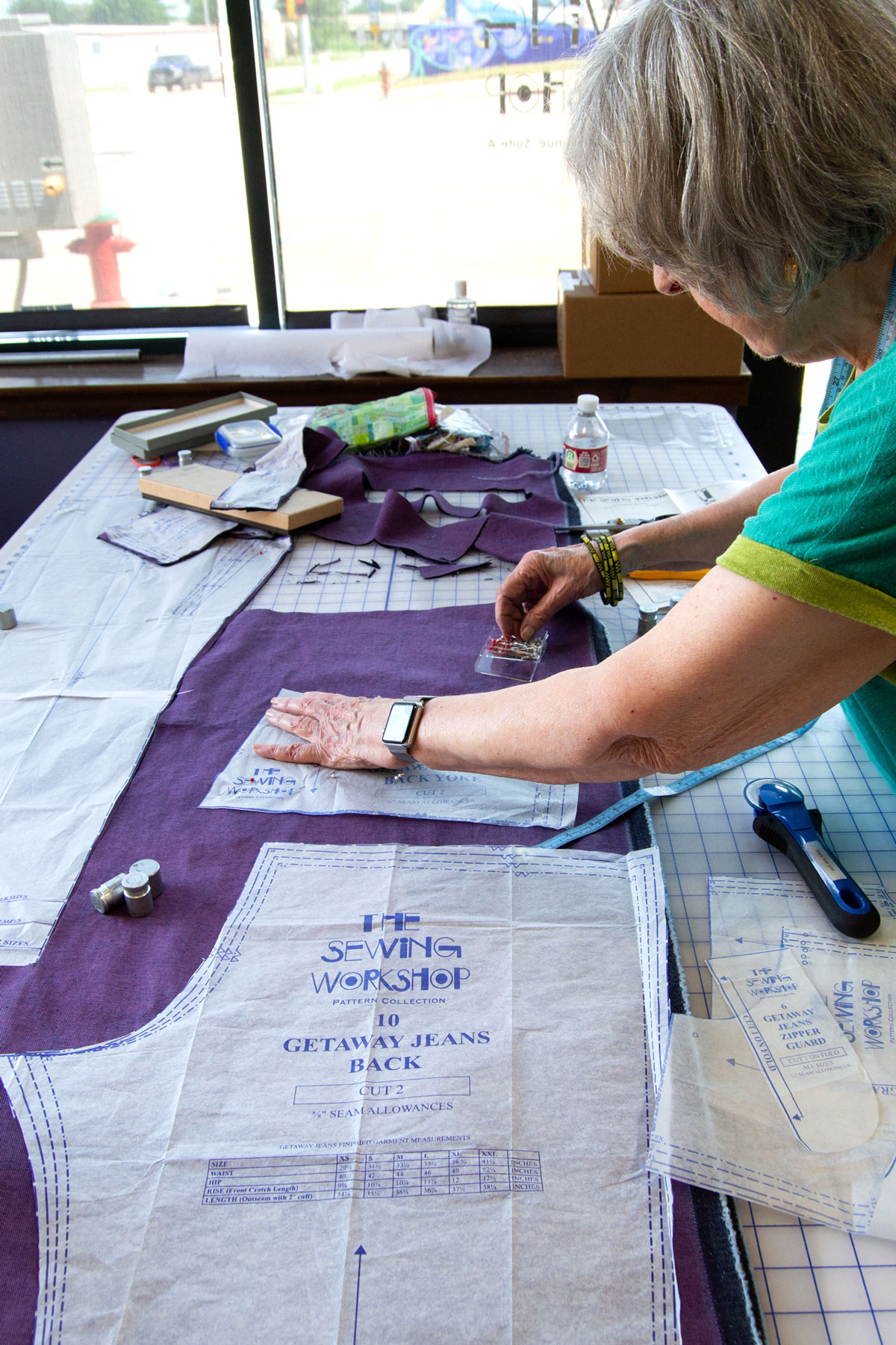 Person working on sewing pattern with fabric and tools on table, window in background.