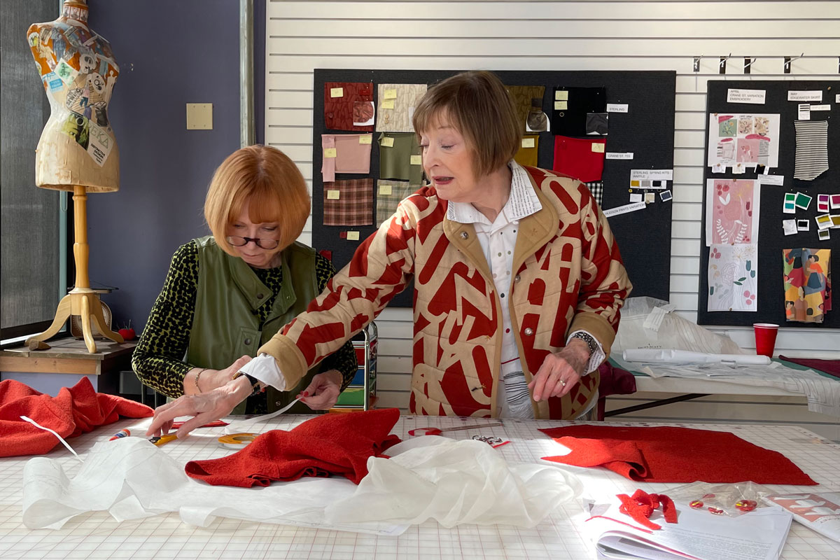 Two women working at a table with red and white fabric, in a room with design boards and a mannequin.