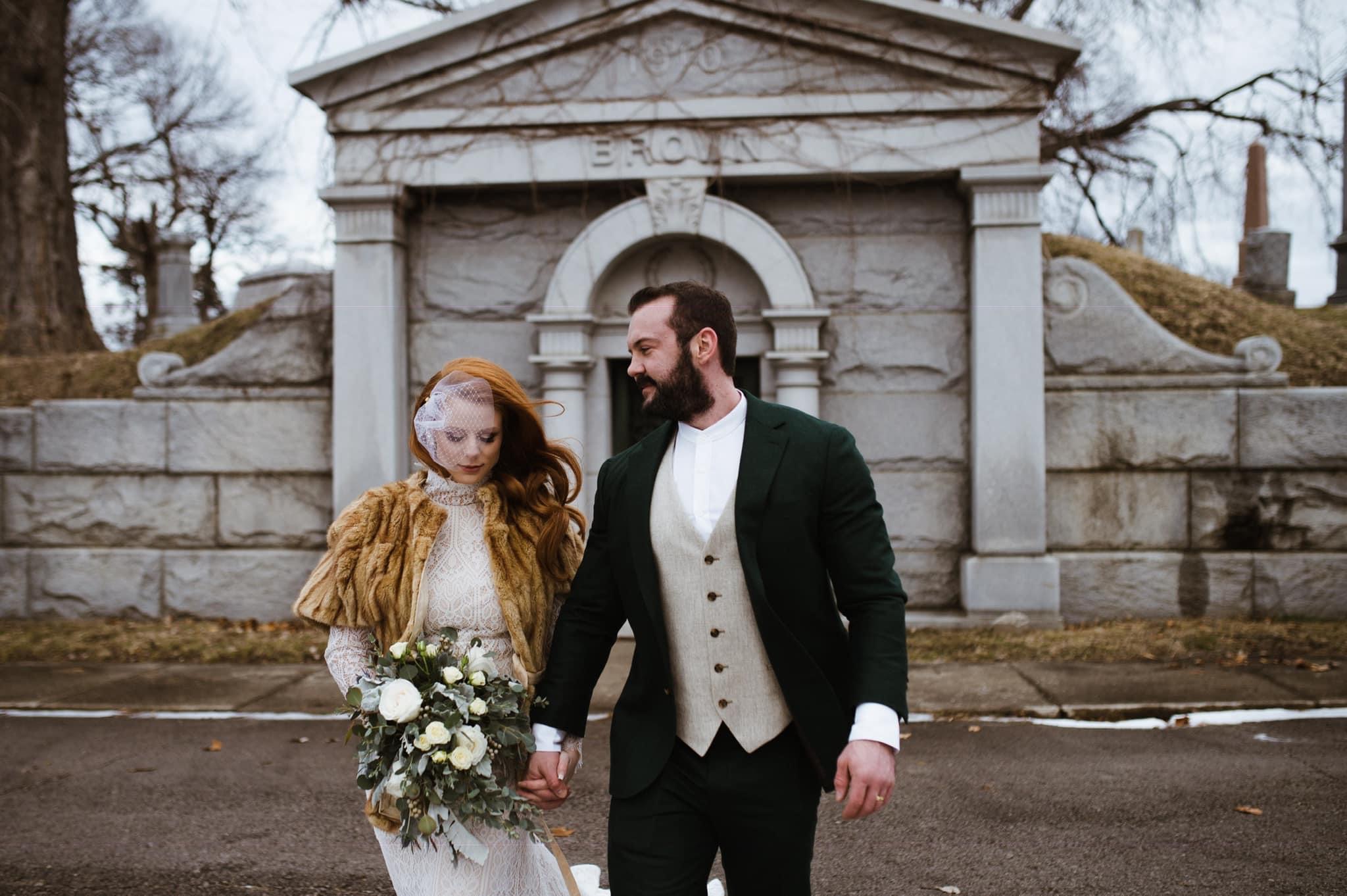A couple holding hands in front of a stone monument, with trees and a cloudy sky in the background.