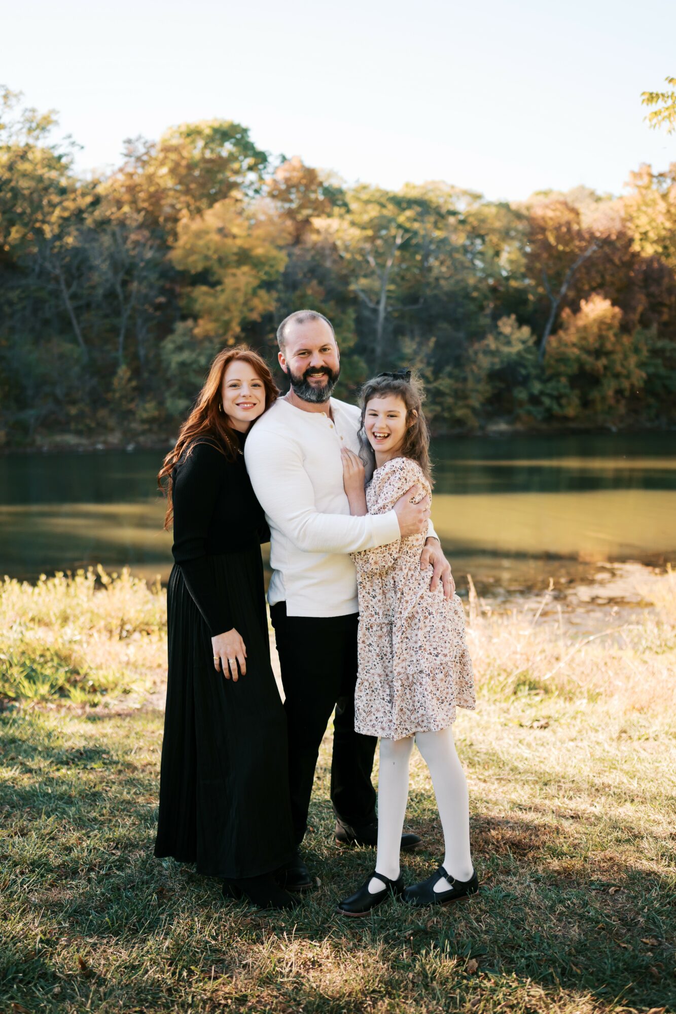 Three people standing outdoors near a body of water with trees in the background, smiling, in sunlight.