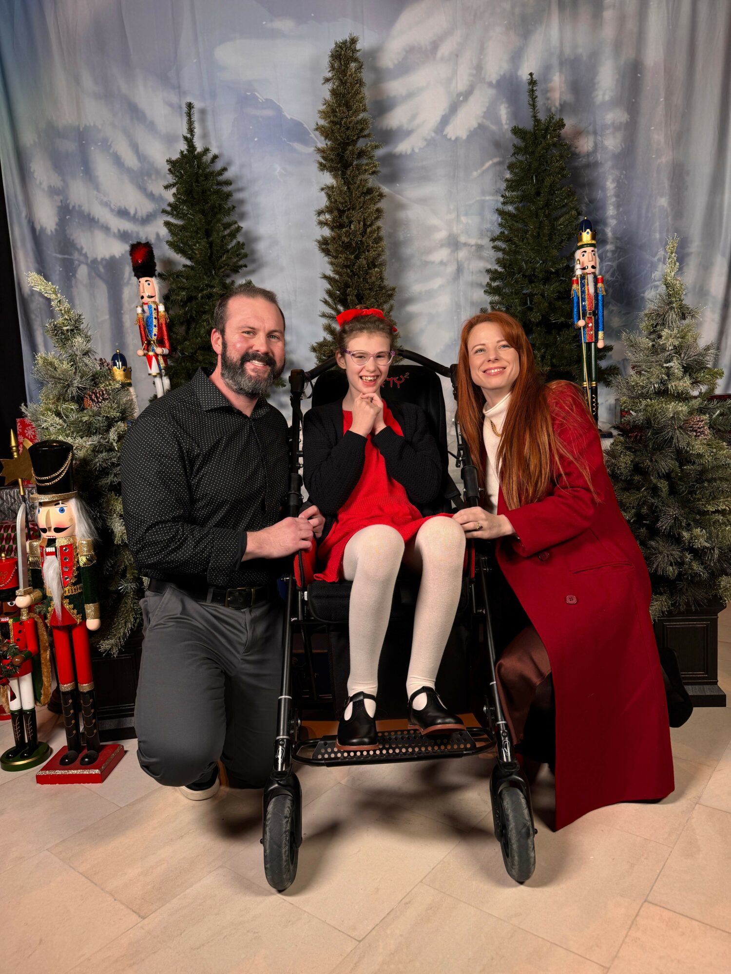 Three people posing with a decorated Christmas tree, two sitting and one standing, in a festive setting.