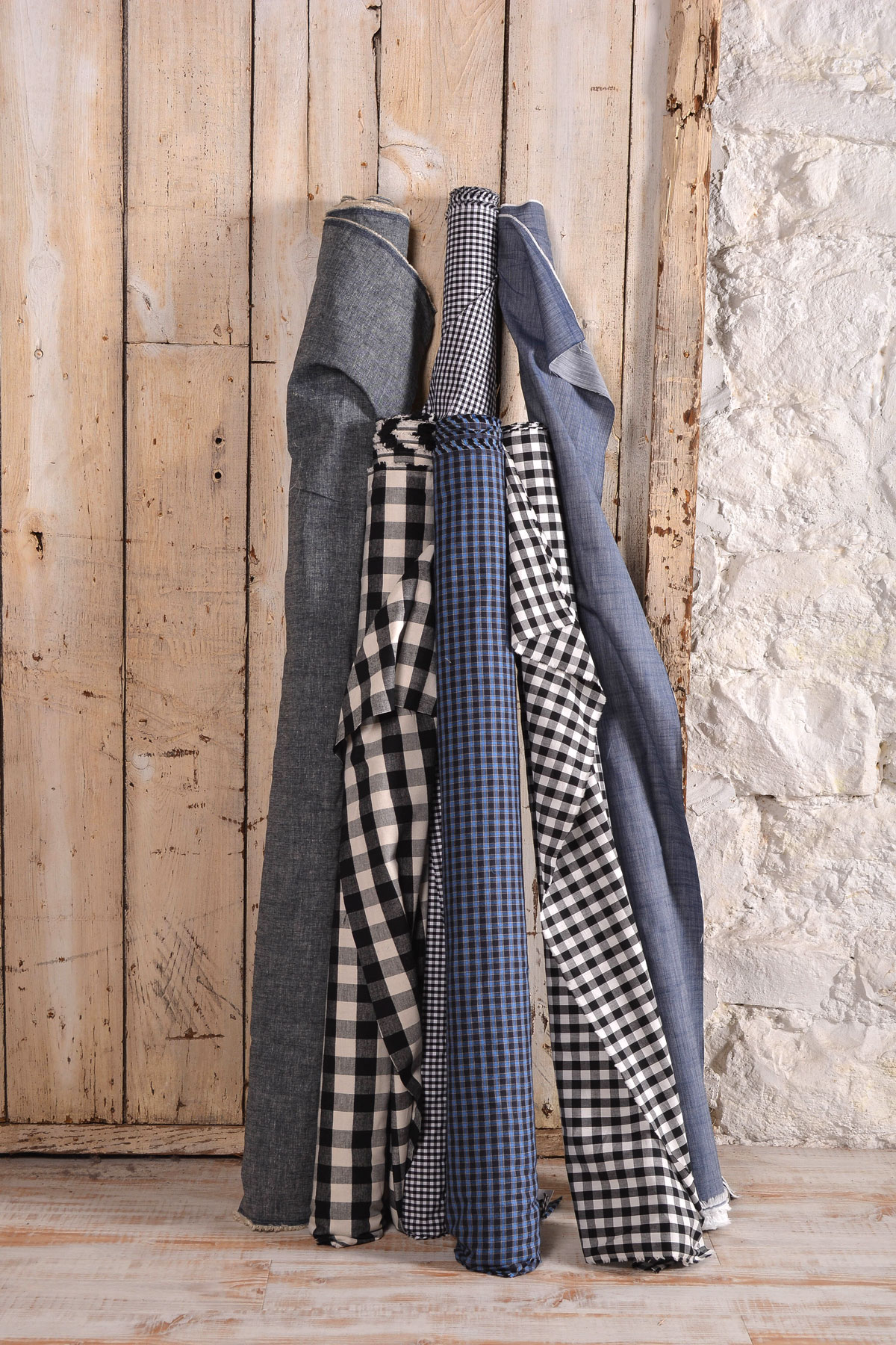 Stacked folded jeans and checkered shirts leaning against a wooden door and stone wall, vertical orientation.