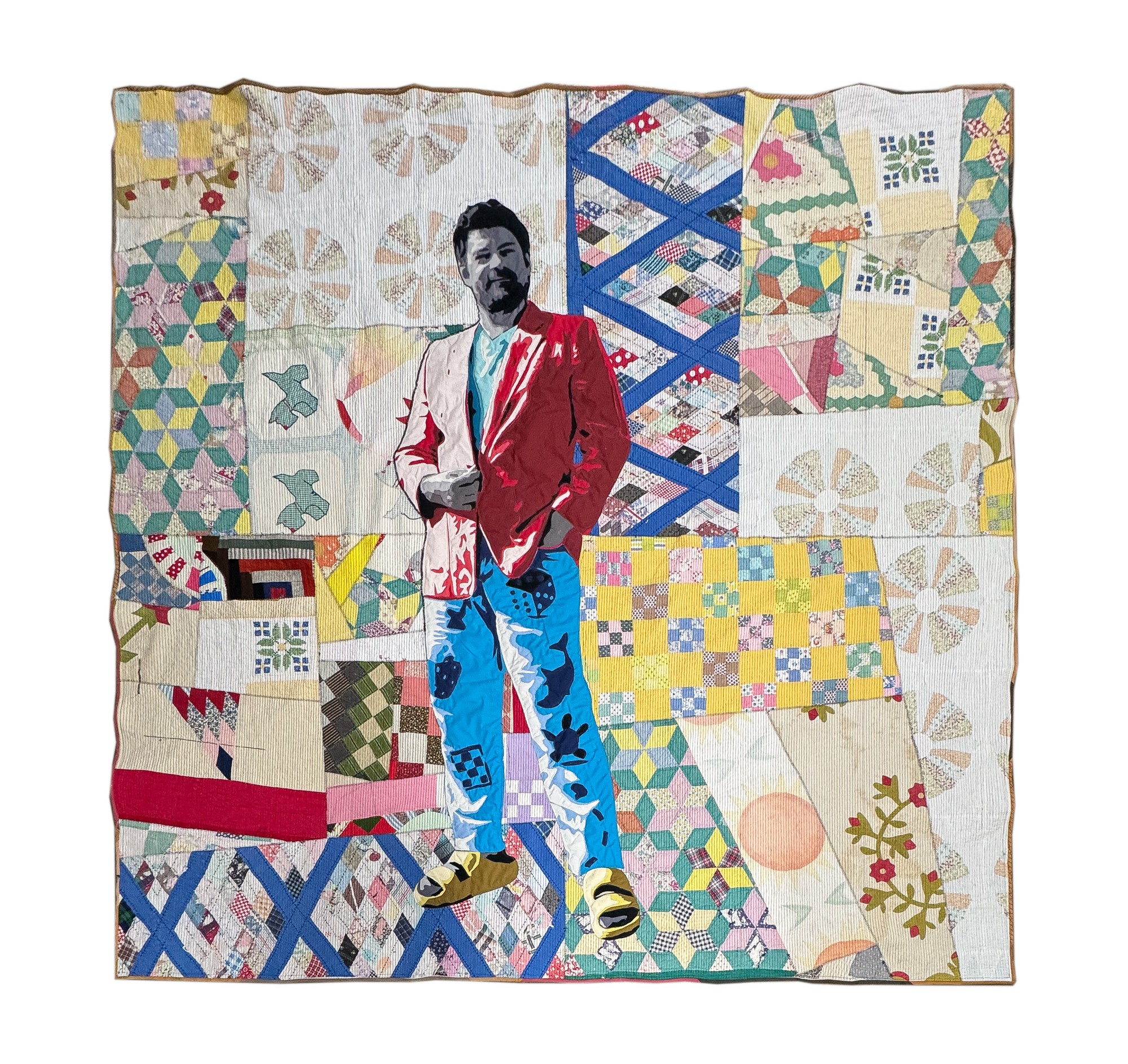 Person standing in front of a colorful patchwork quilt, wearing a red jacket and blue pants, with a beard and mustache.