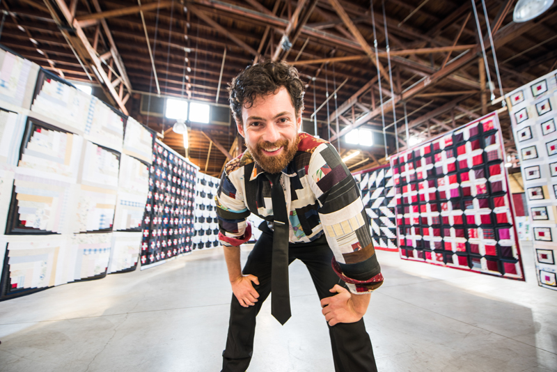 Man with a beard and curly hair smiling, leaning forward in a store aisle with fabric rolls on shelves behind him.