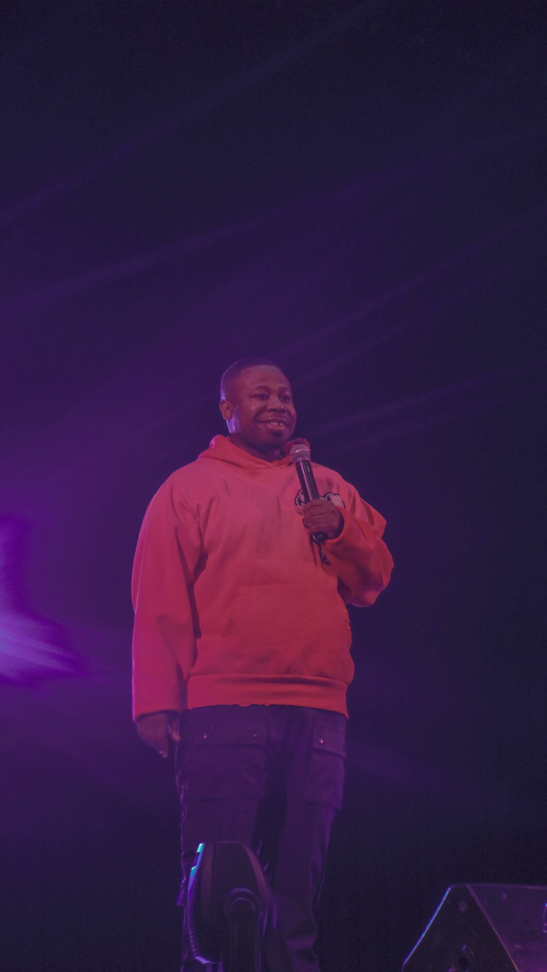 Person standing on stage holding microphone, wearing red hoodie, dark pants, smiling, dark background with stage lighting.