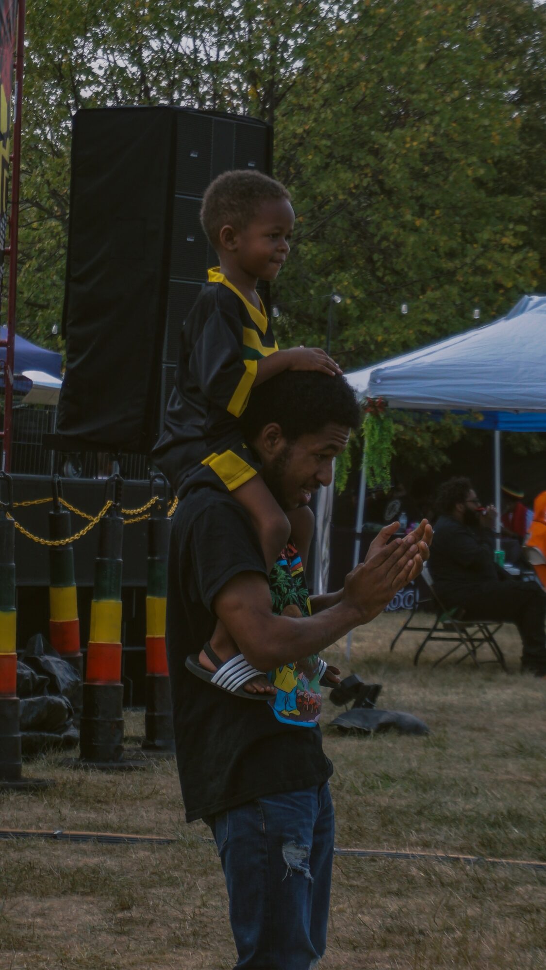 Two children in black shirts with yellow accents outdoors, one clapping and the other standing near a speaker and tent.