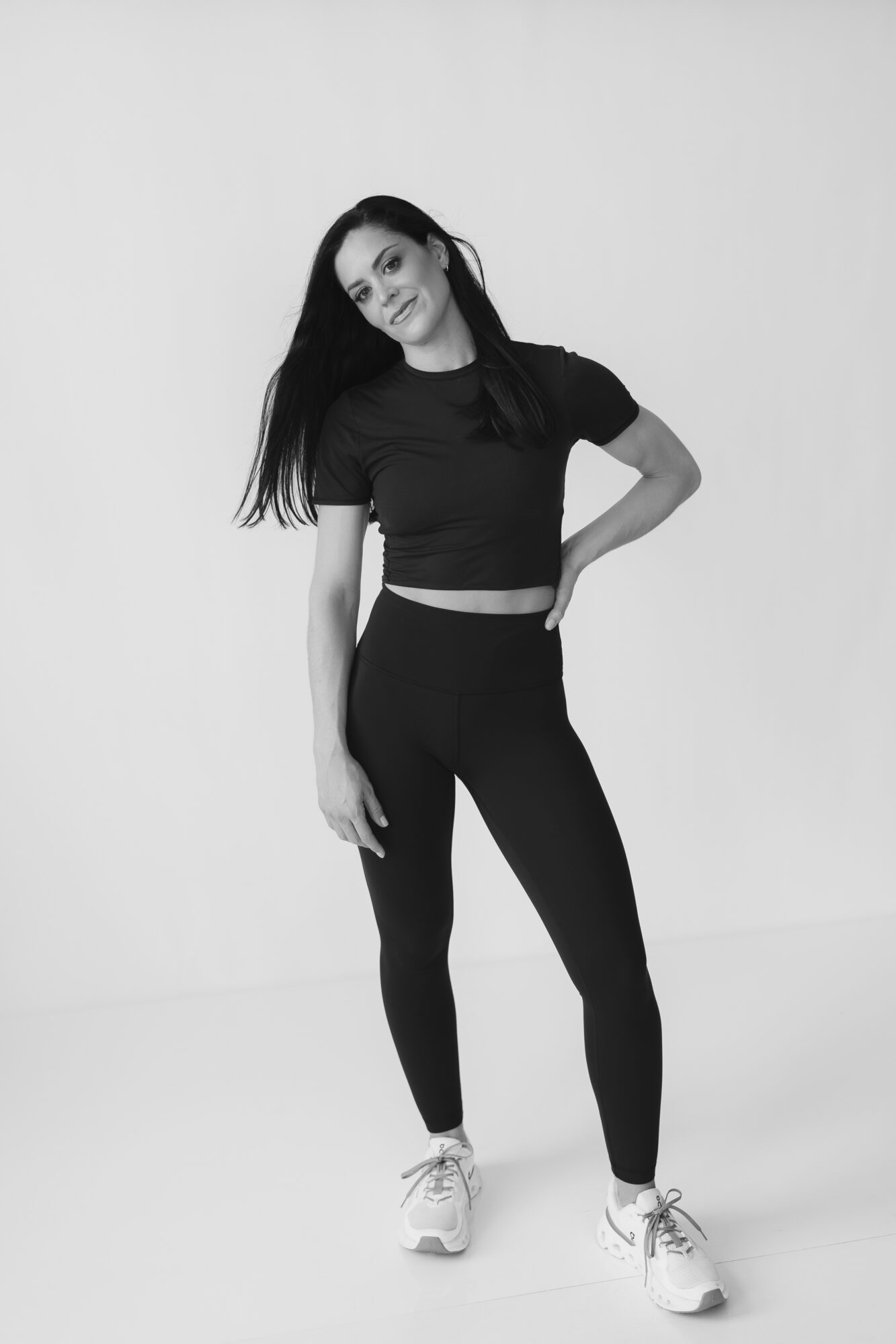 Young woman with long hair in black crop top and leggings, standing against plain background, smiling slightly, hands on hips, facing camera.