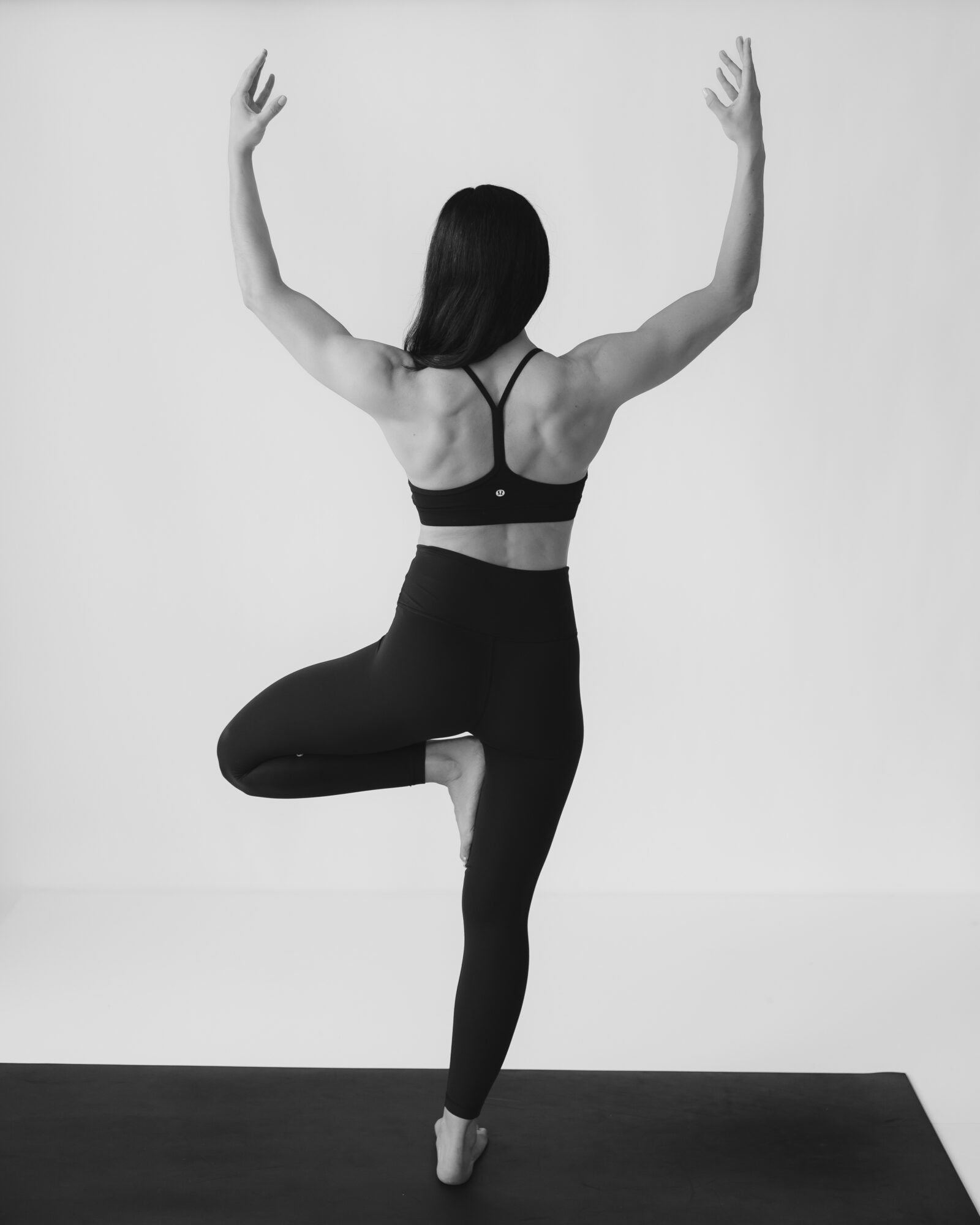 Person practicing yoga in tree pose with arms raised, facing away, against a plain wall, on a mat.