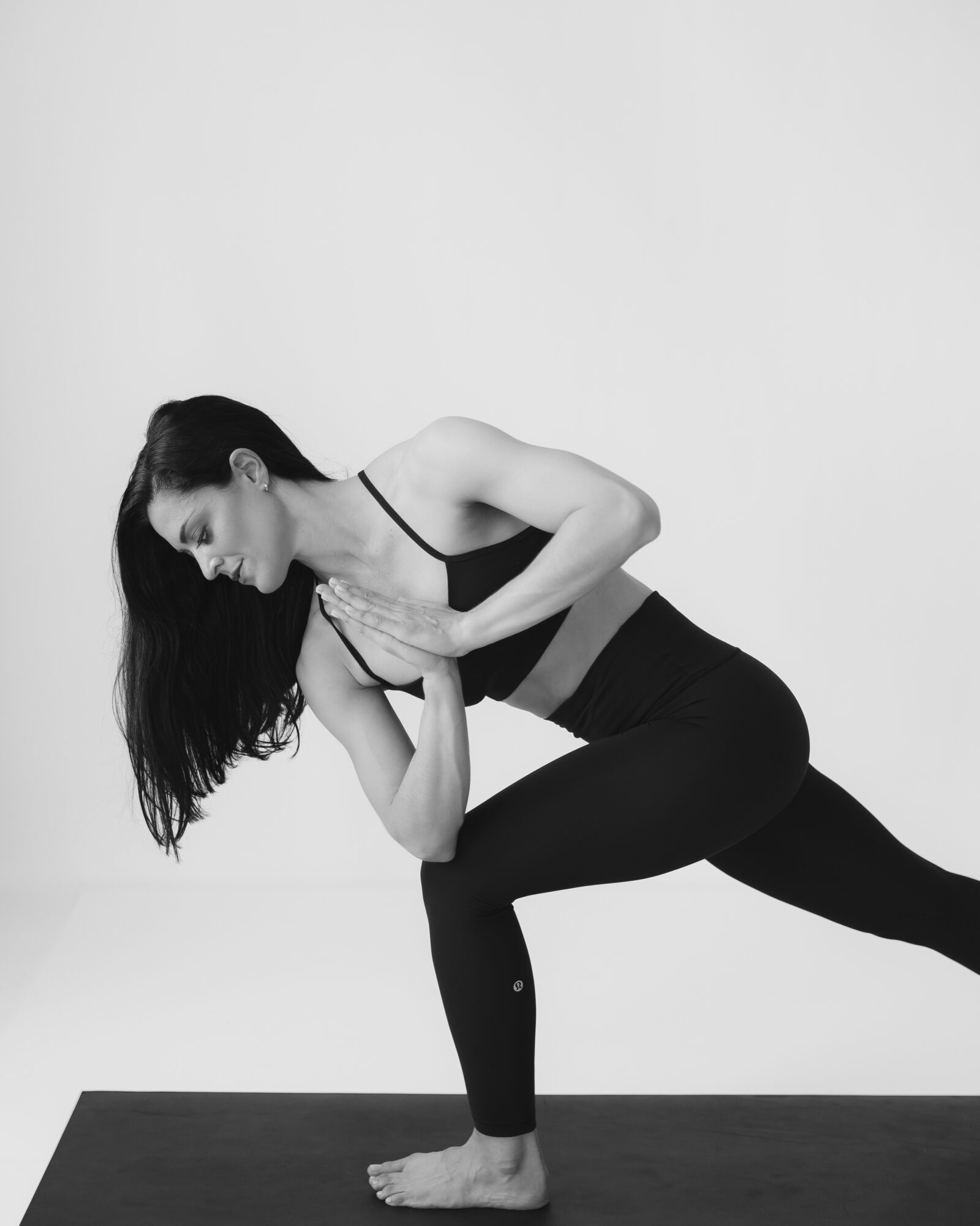 Woman practicing yoga in a lunge position, hands pressed together in prayer, on a mat against a plain background.