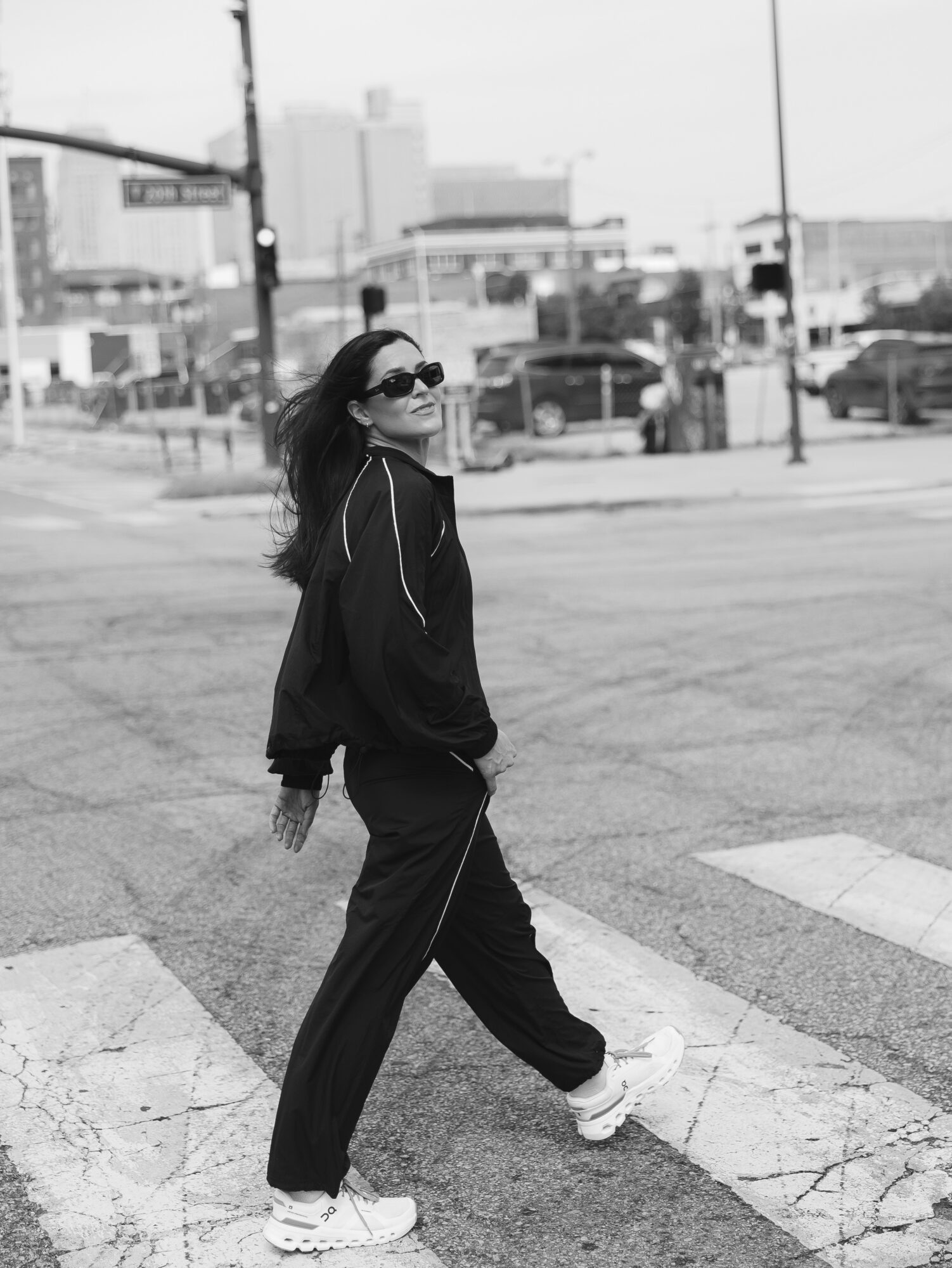 Woman walking across a crosswalk in an urban area, wearing sunglasses and casual clothing, with city buildings in background.