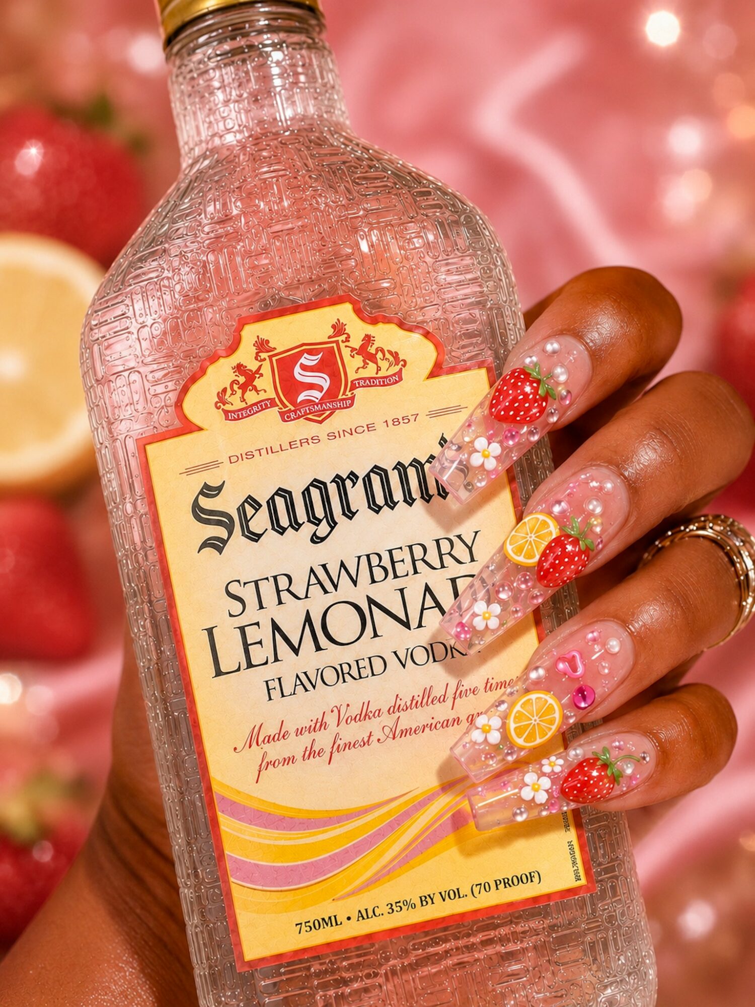 Hand with decorated nails holding a Seagram's Strawberry Lemonade bottle, background with strawberries and lemon, pink backdrop.