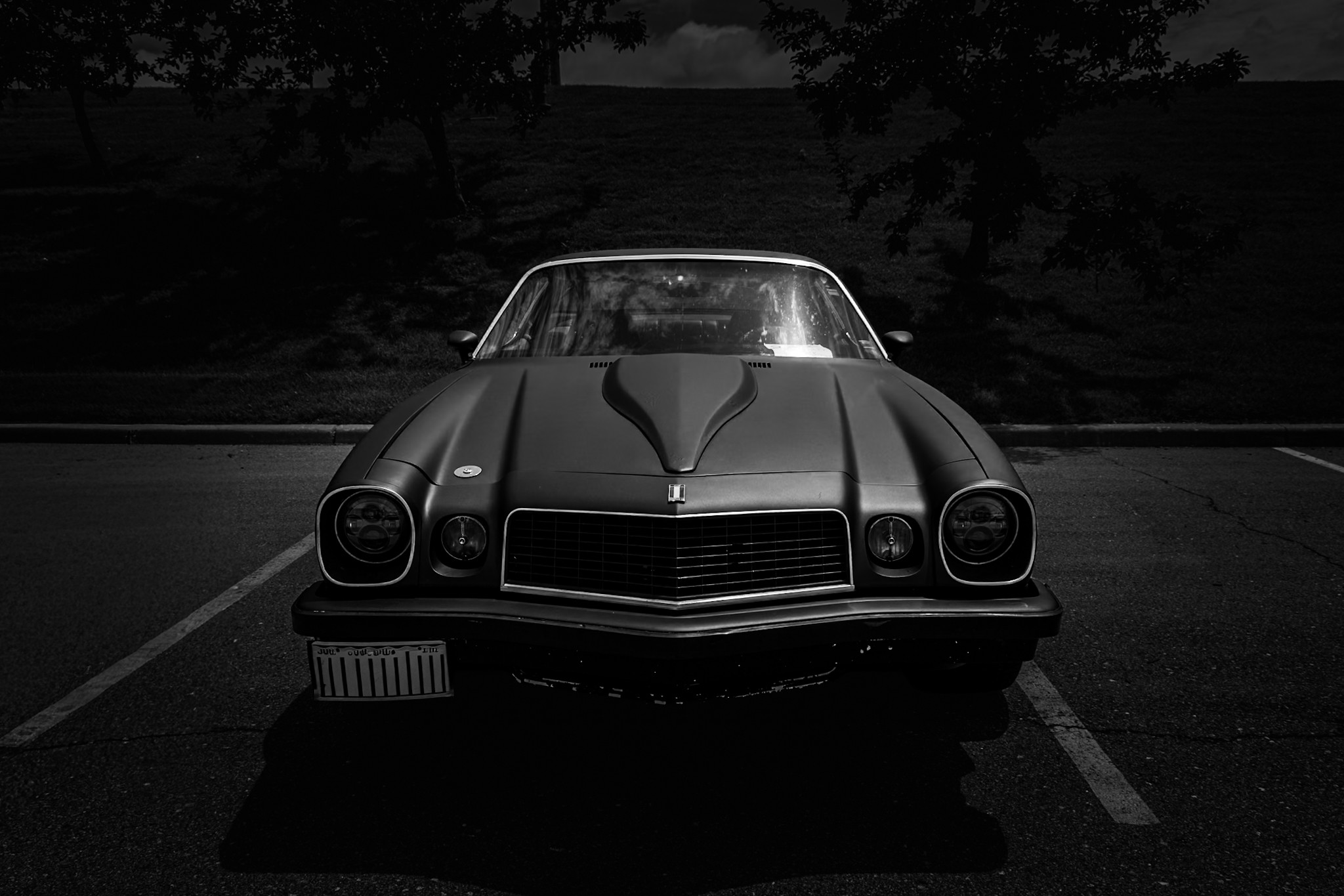 Front view of a vintage car parked in a lot, with trees and a building in the background, in black and white.