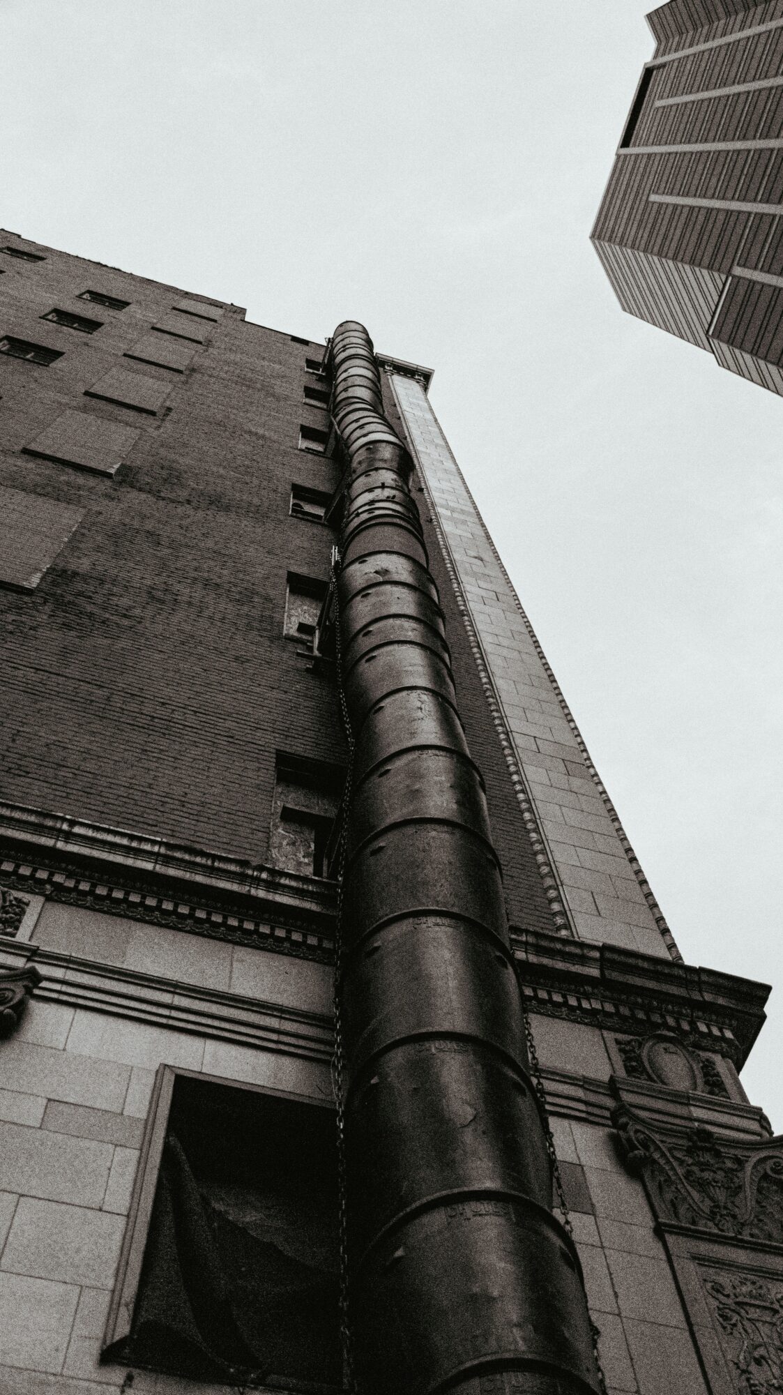 View of tall buildings from ground looking up, with a large pipe running down the side of a building.
