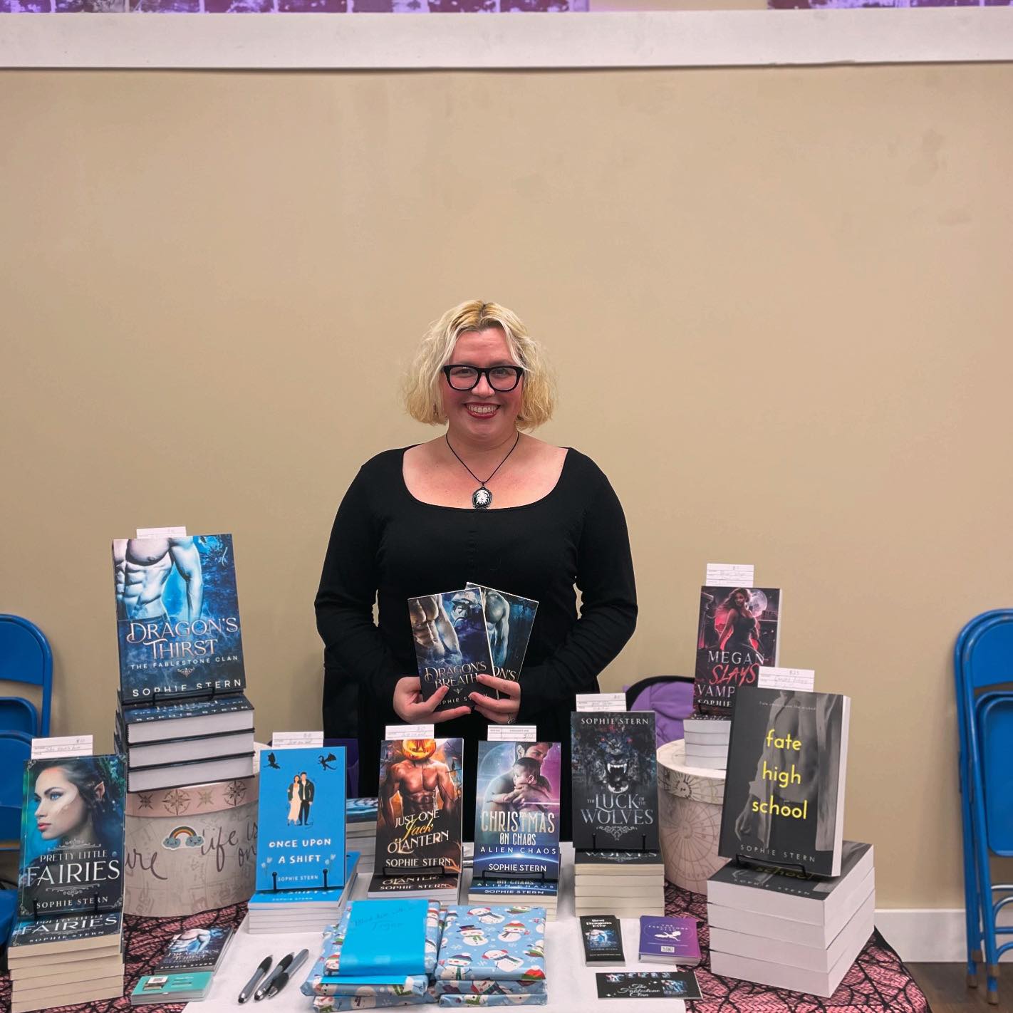 Woman with glasses and blonde hair standing behind a table of books, smiling, in a room with blue chairs.