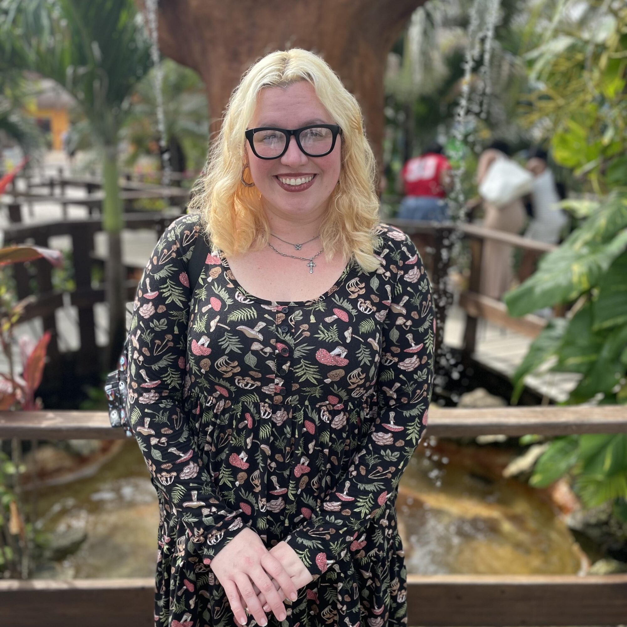Smiling woman with blonde hair, glasses, wearing a floral dress, standing outdoors with greenery and a pond behind her.