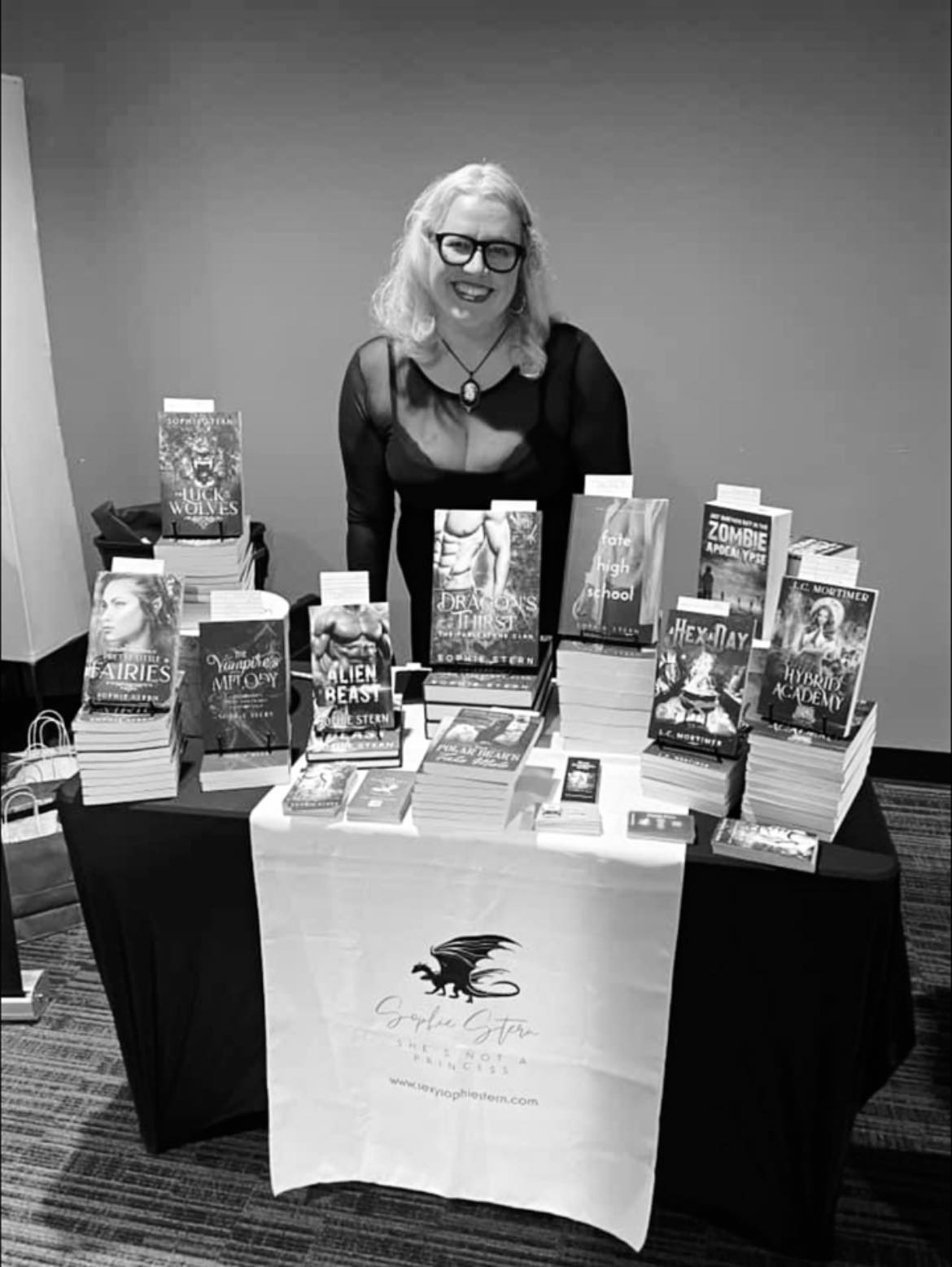 Woman with glasses and light hair standing behind a table with books, smiling, in a room with plain wall.