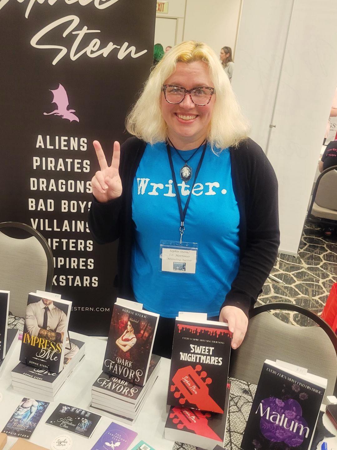 Woman with blonde hair and glasses smiling at a table with books, making a peace sign, wearing a blue shirt with 'Writer.'