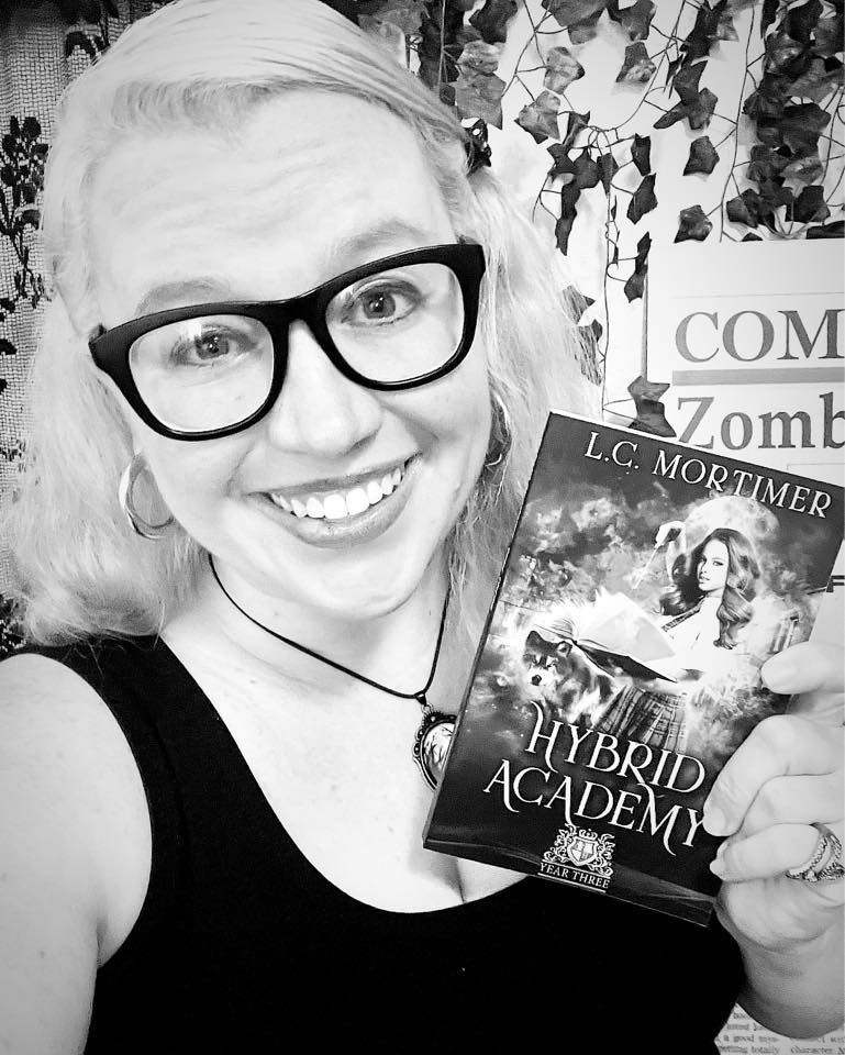 Woman with glasses smiling, holding a book titled 'Hybrid Academy' in front of a leafy background.