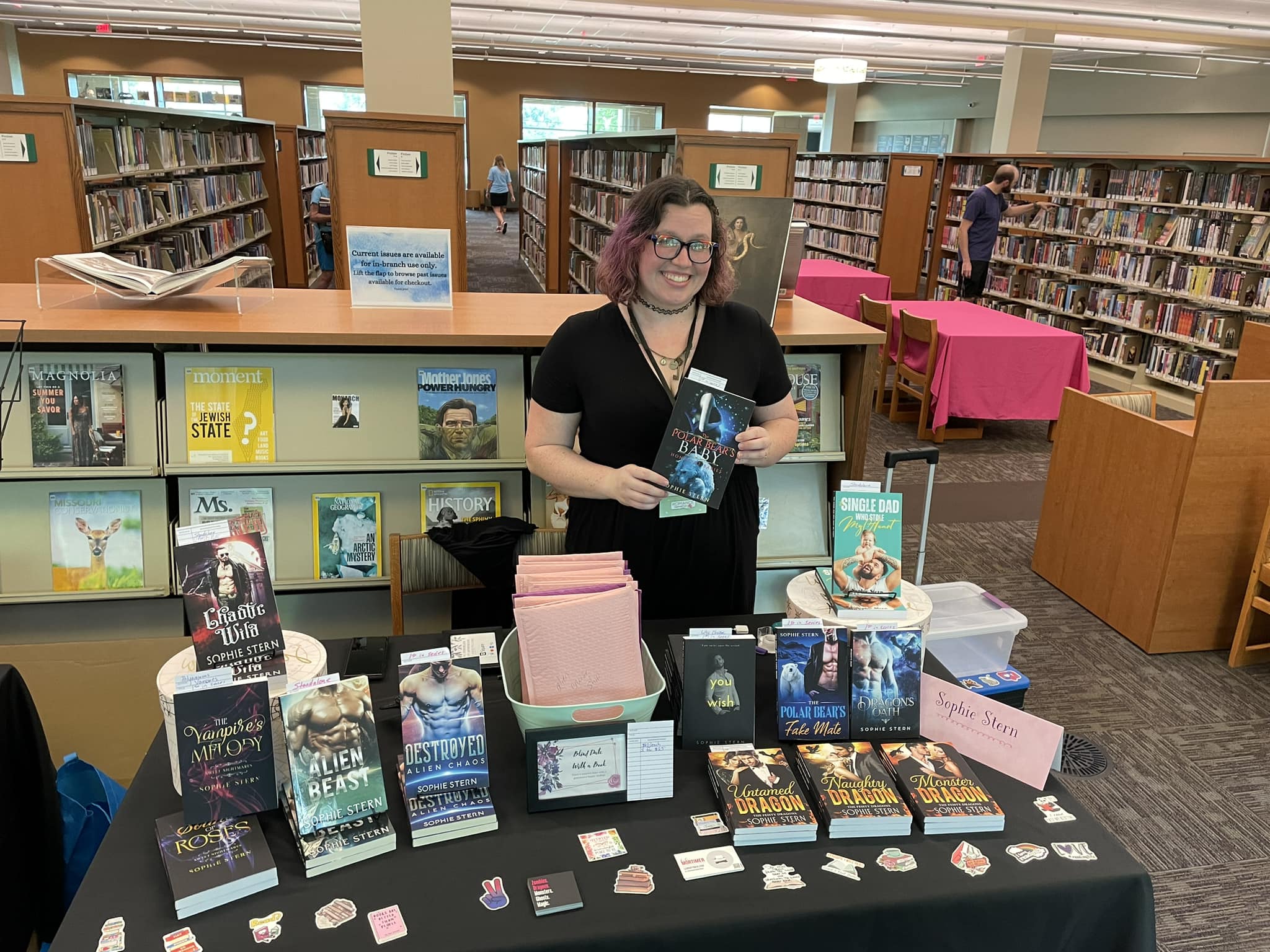Woman standing behind a table of books in a library, holding a book, with bookshelves in the background.