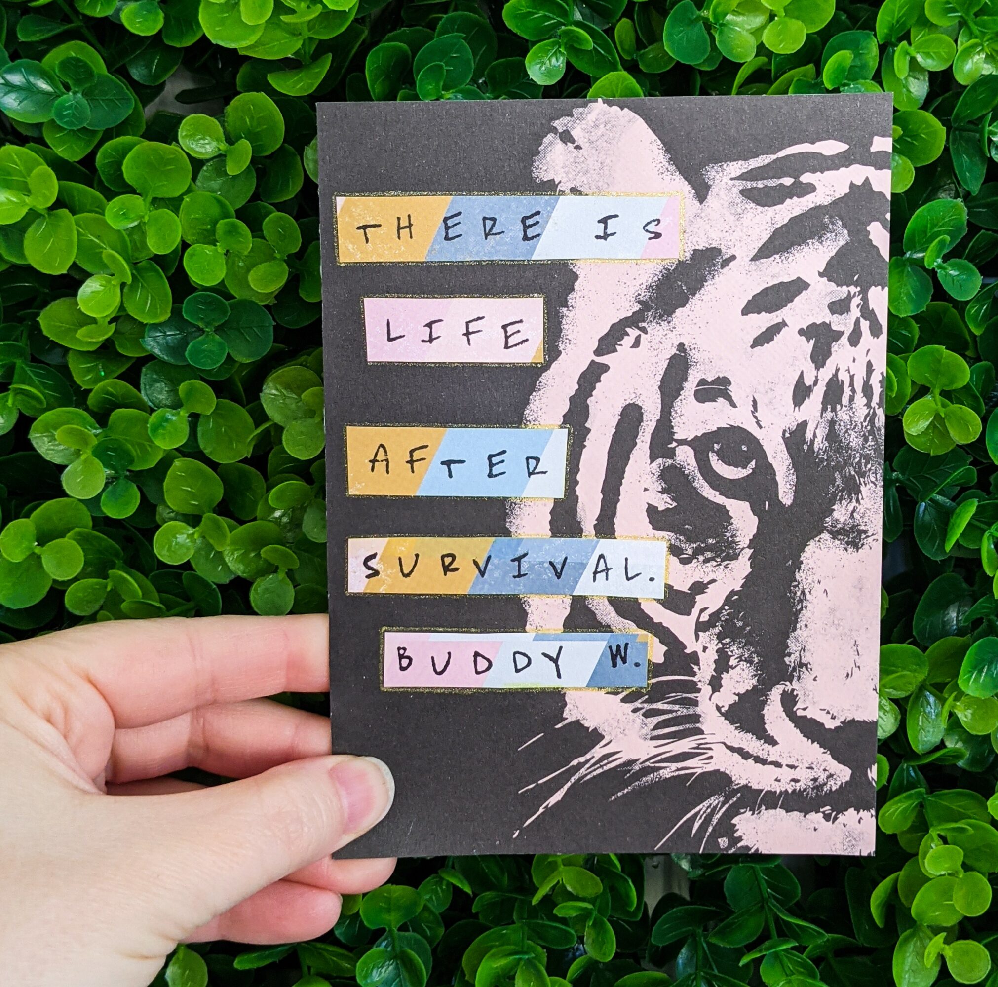 Hand holding a card with a tiger illustration and colorful text, against green foliage background.