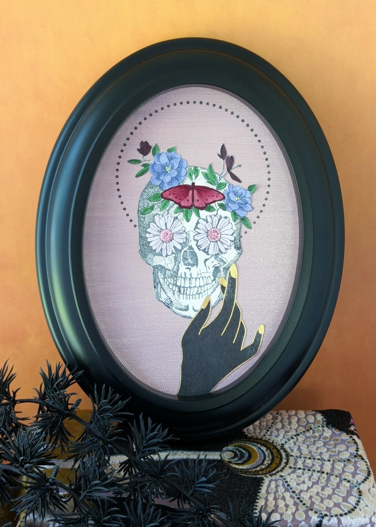 Framed artwork of a skull with flowers, a butterfly, and a hand with painted nails, against a peach background.