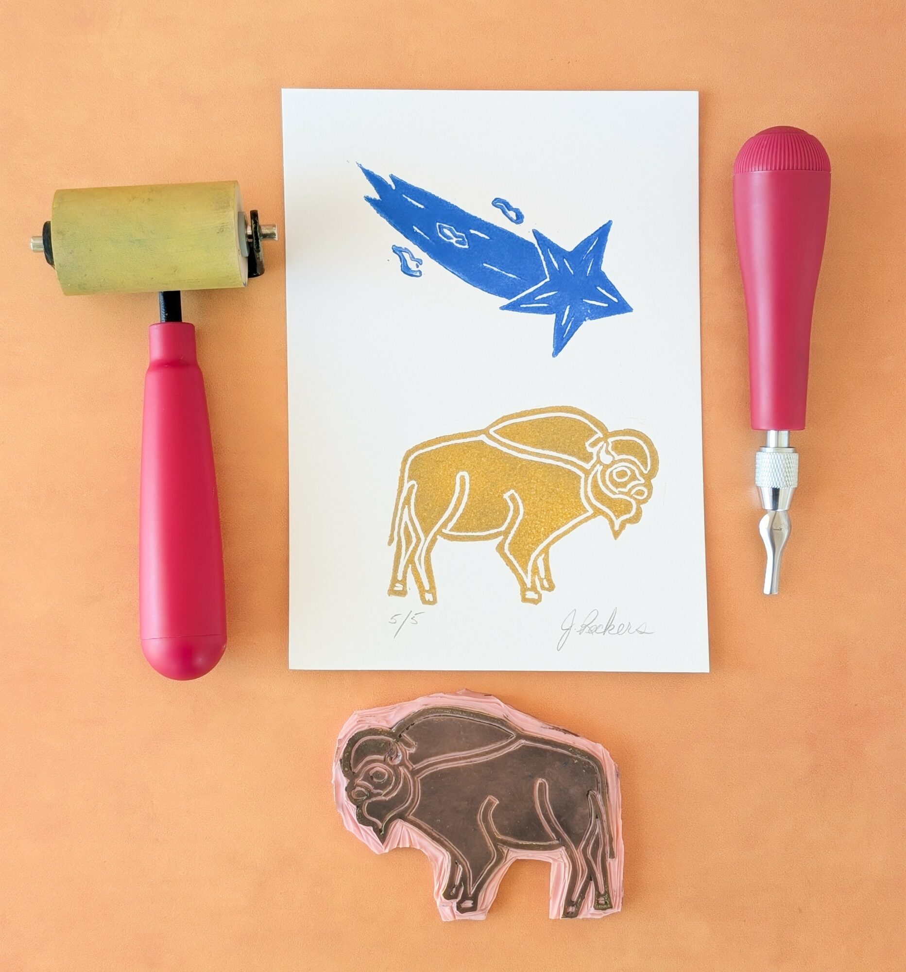 Prints of a blue shooting star and a yellow buffalo on paper, with a pink roller and a pink-handled tool nearby.