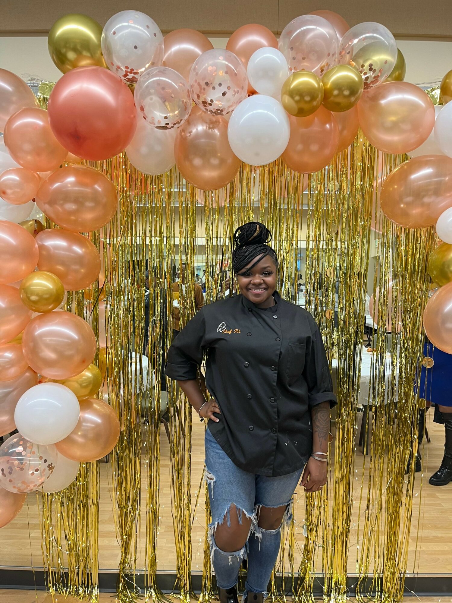 Person standing in front of a balloon and gold curtain backdrop, wearing a black jacket and jeans, with hands on hips.