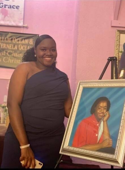 Smiling woman in a dark dress standing next to a framed portrait of a person in a red shirt, pink wall background.