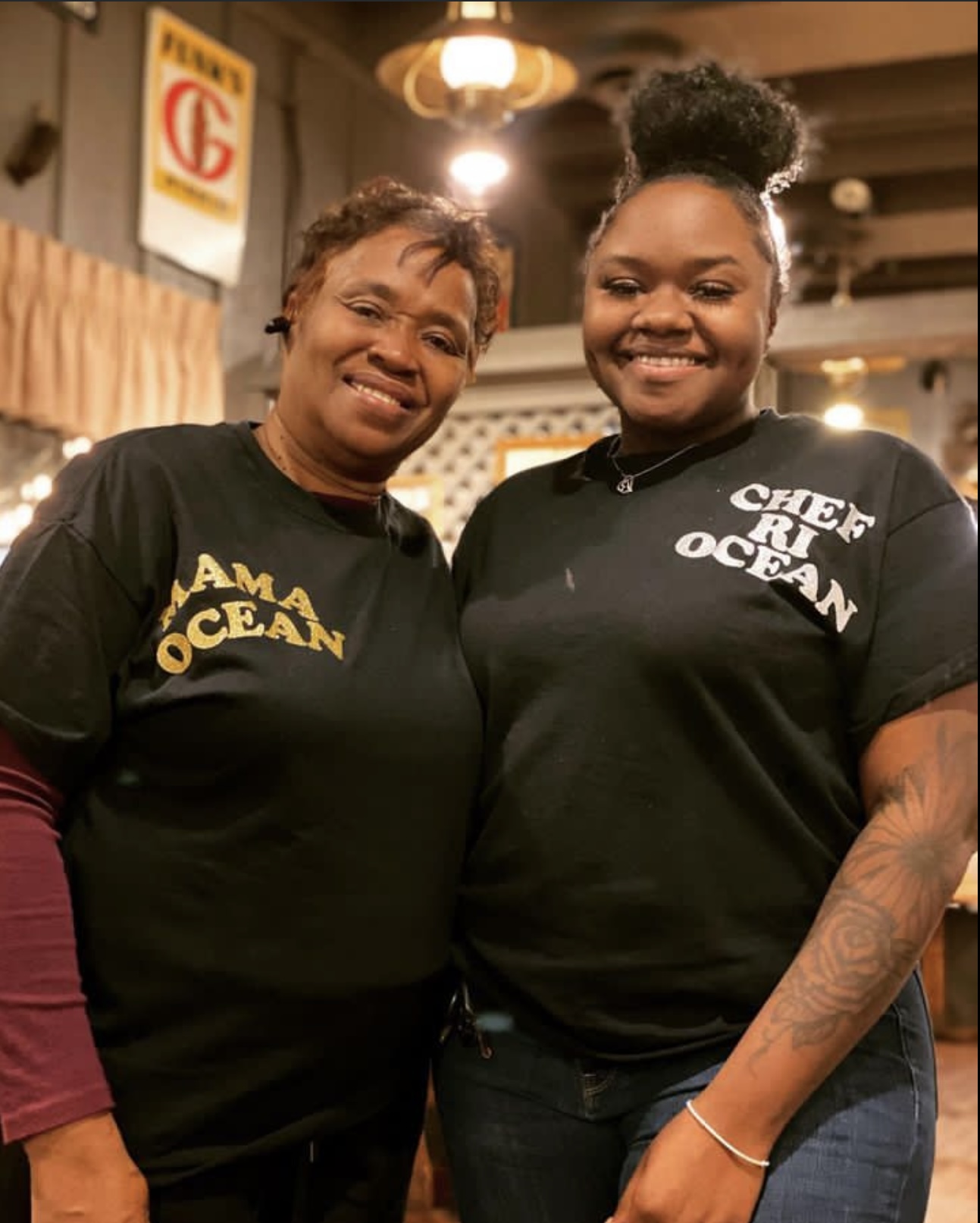 Two women smiling, standing indoors with warm lighting, one wearing a black shirt with yellow text, the other with a tattooed arm and a black shirt with white text.