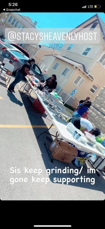 People gather around tables outdoors with cars and buildings in background, some supporting or grinding, in a street scene.