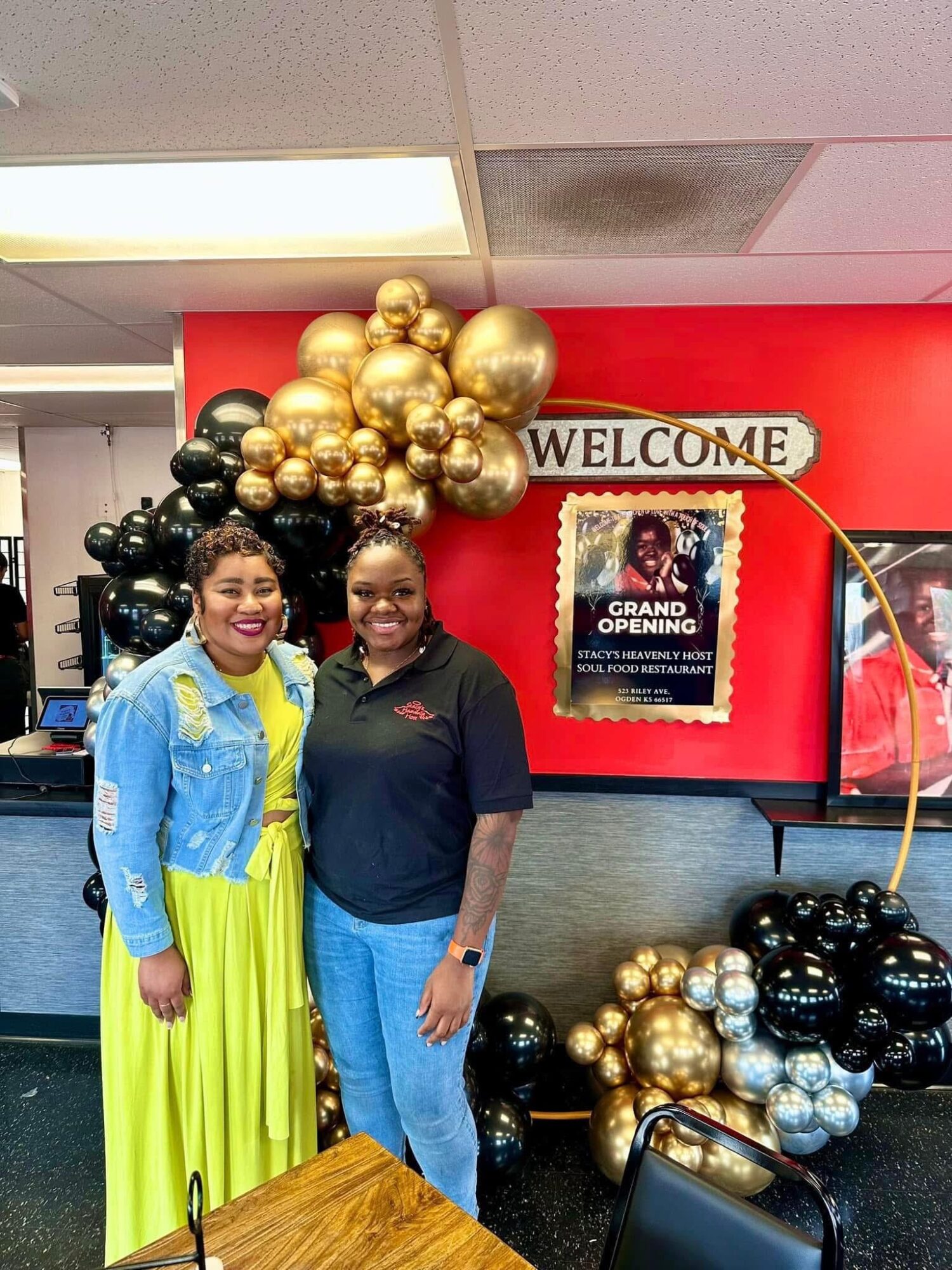 Two women stand in front of a red wall decorated with gold, black, and silver balloons, and a welcome sign, inside a building.