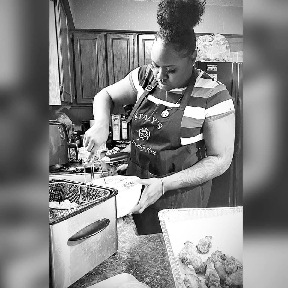 Woman with curly hair in a striped shirt and apron preparing food in a kitchen, with fried items on a tray nearby.