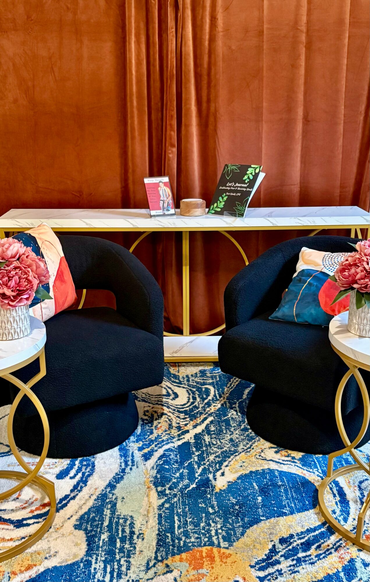 Two black armchairs with colorful pillows, gold side tables with flowers, and a blue patterned rug in front of a wooden wall.