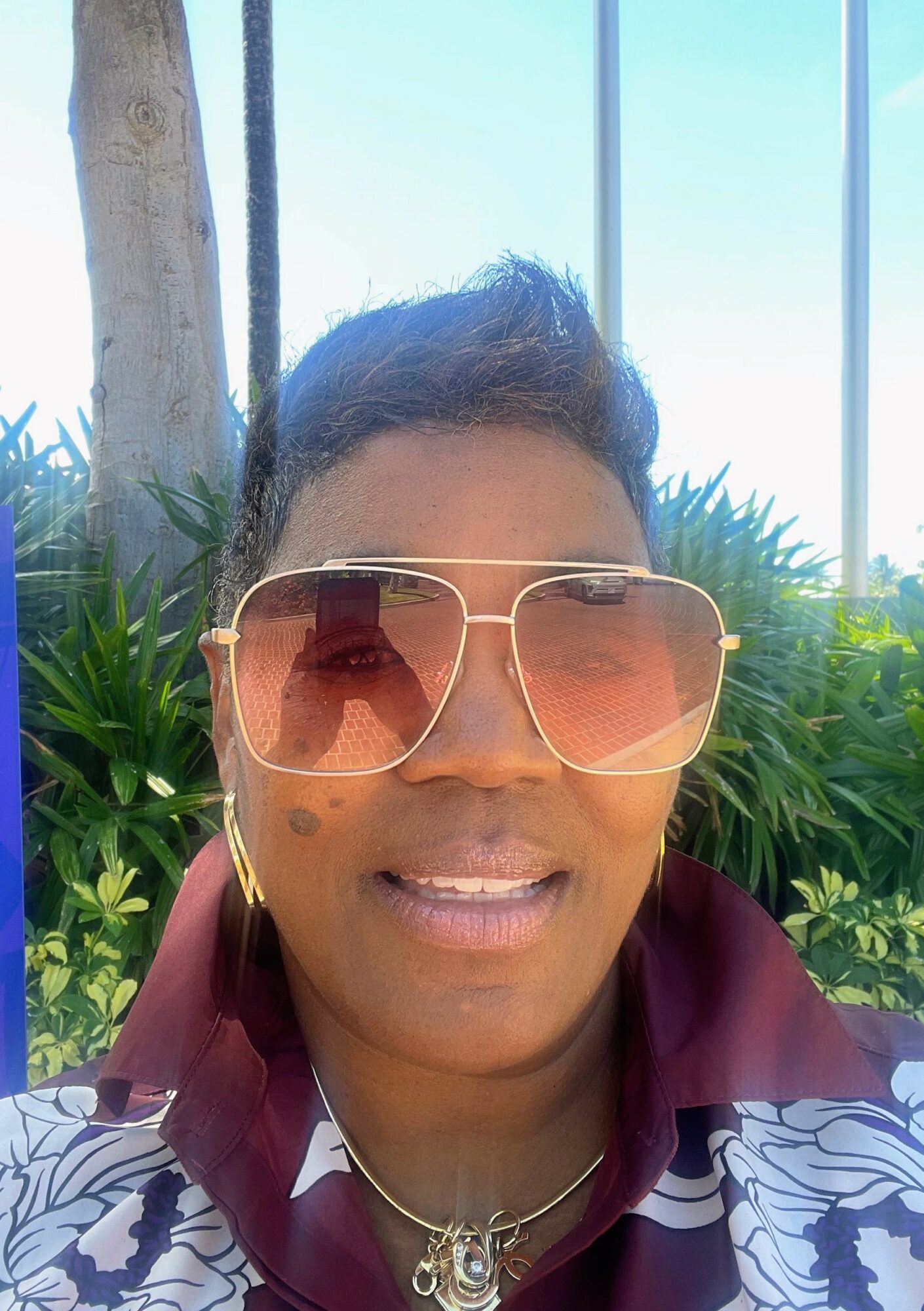 Person wearing large sunglasses and a maroon shirt outdoors with greenery and blue sky behind.