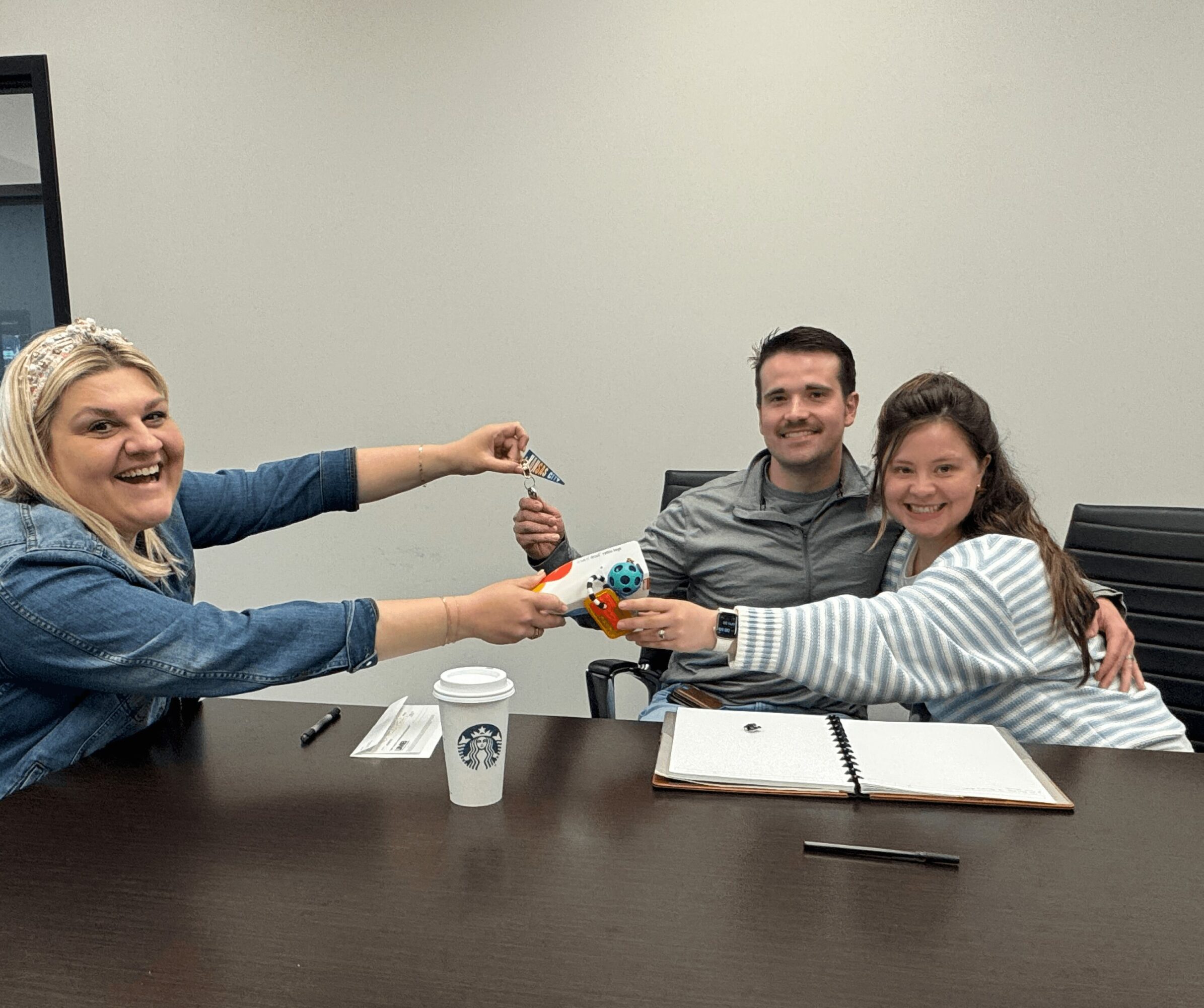 Three people sitting at a table exchanging a small toy, smiling, with a Starbucks cup and notebooks on the table.