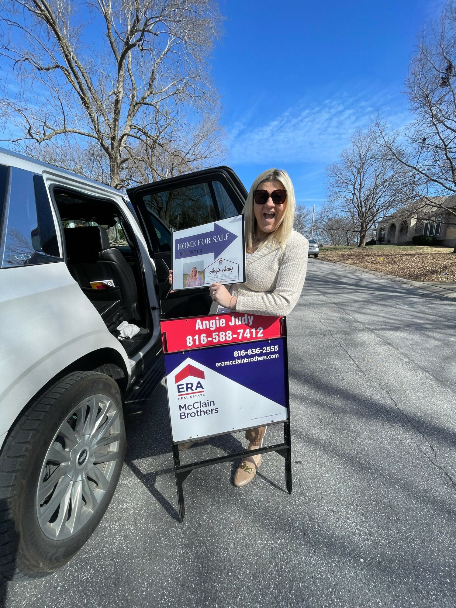 Woman standing next to a car with a signboard on a sunny day, trees and blue sky in background.