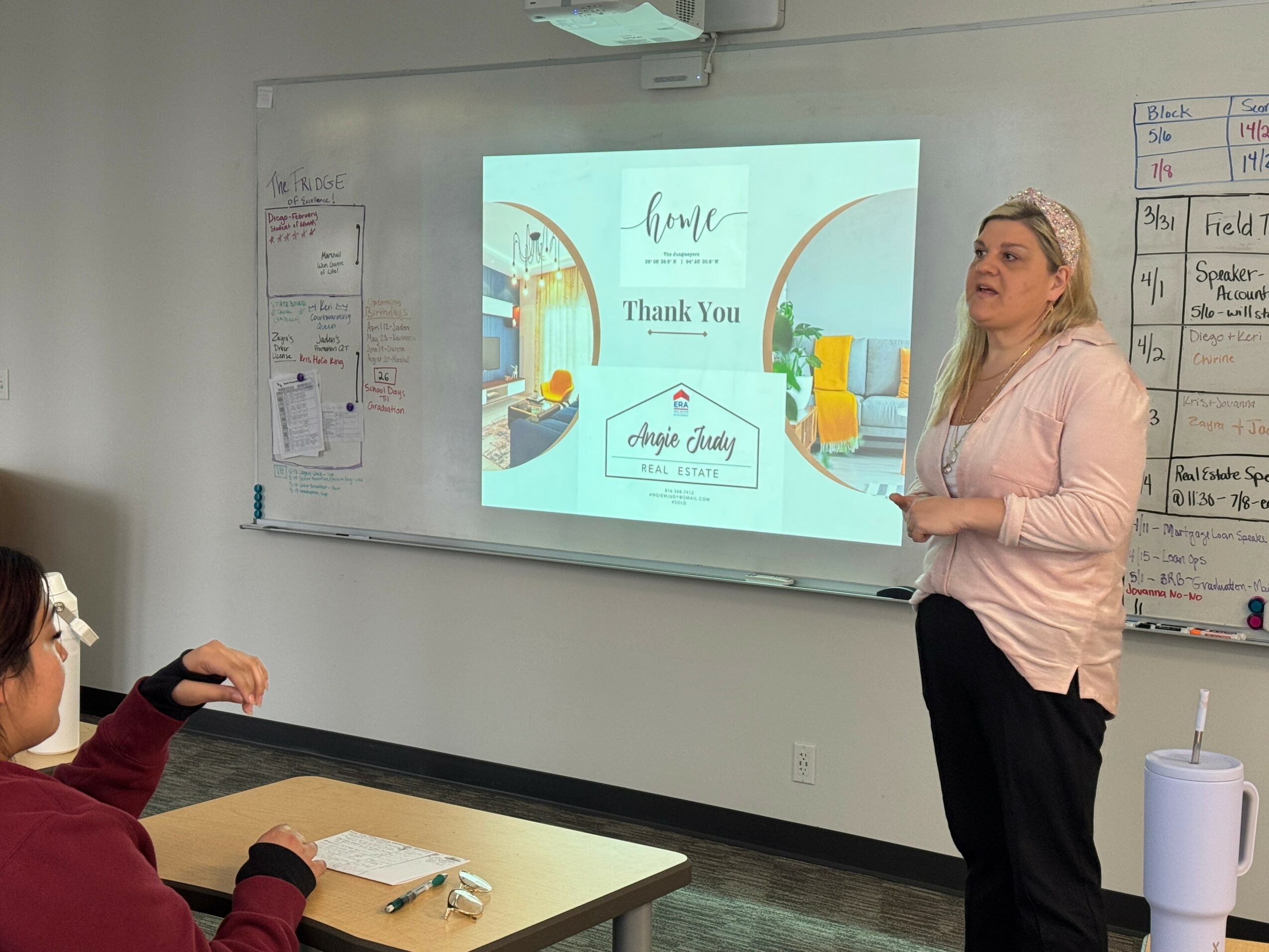 Woman standing in front of a classroom whiteboard giving a presentation, with students seated at desks. Projected slide says 'Thank You' and 'Enjoy Your Day'.