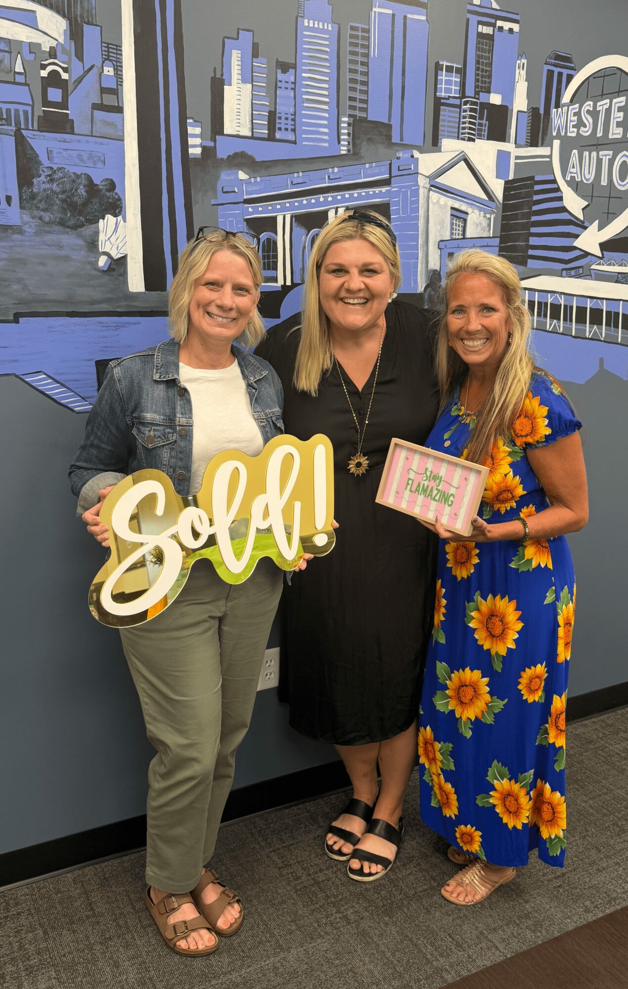Three women standing together indoors, smiling, with a cityscape mural in the background. One holds a sign that says 'Eola!'.