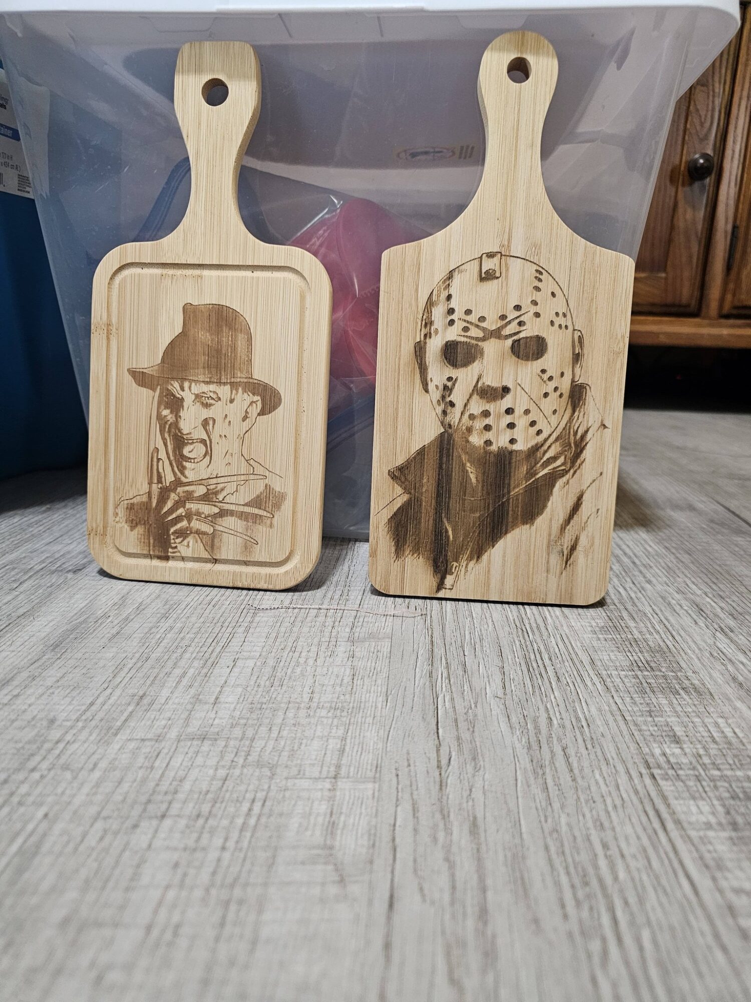 Two wooden cutting boards with engraved images of horror movie characters, one with a hat and the other with a hockey mask, on a wooden floor.