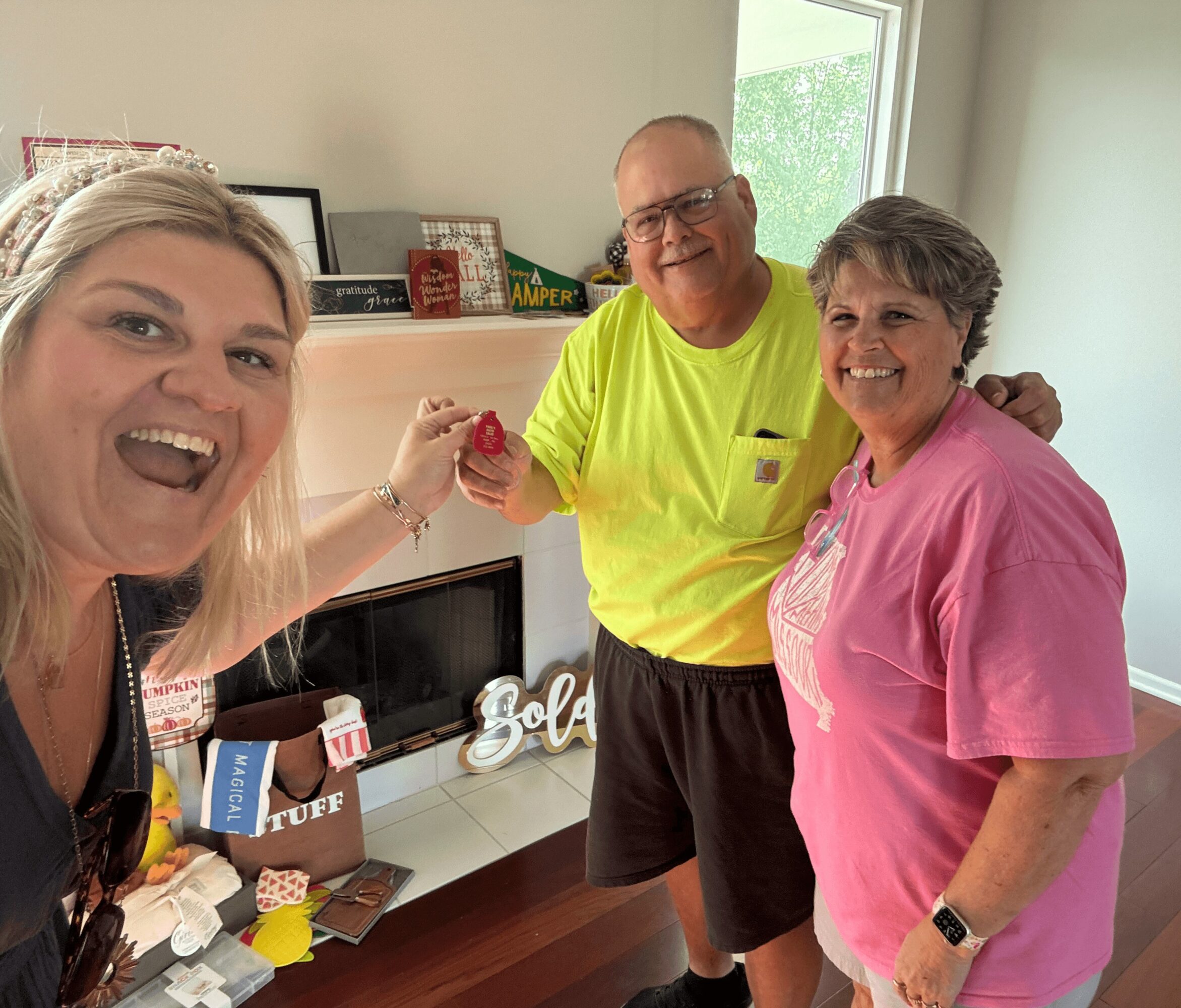 Three people smiling indoors, one holding a red object, with a TV and shelves in the background.