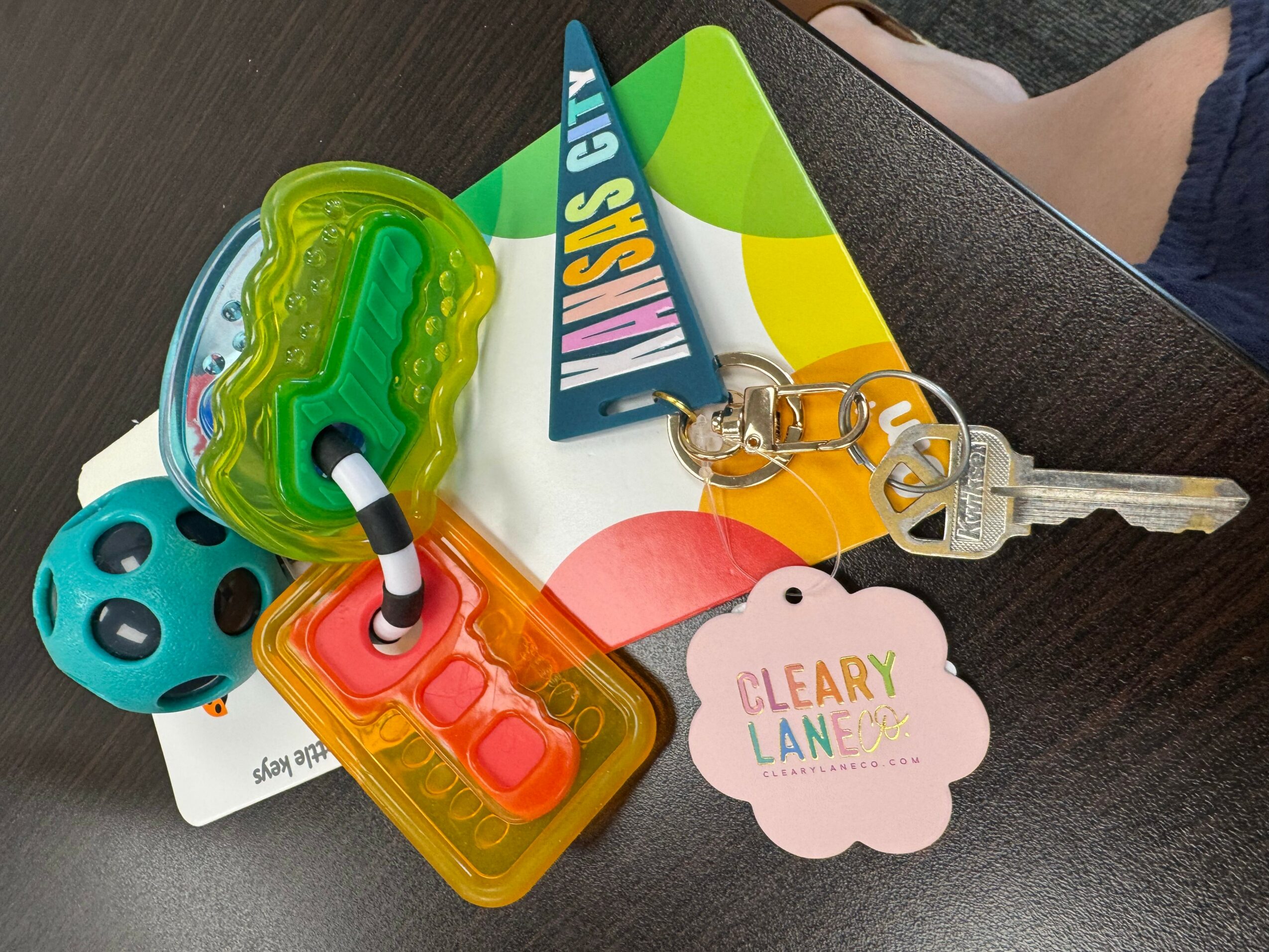 Colorful keychain with a green dinosaur, blue ball, pink and orange popsicle, and a pink cloud-shaped tag on a dark surface.