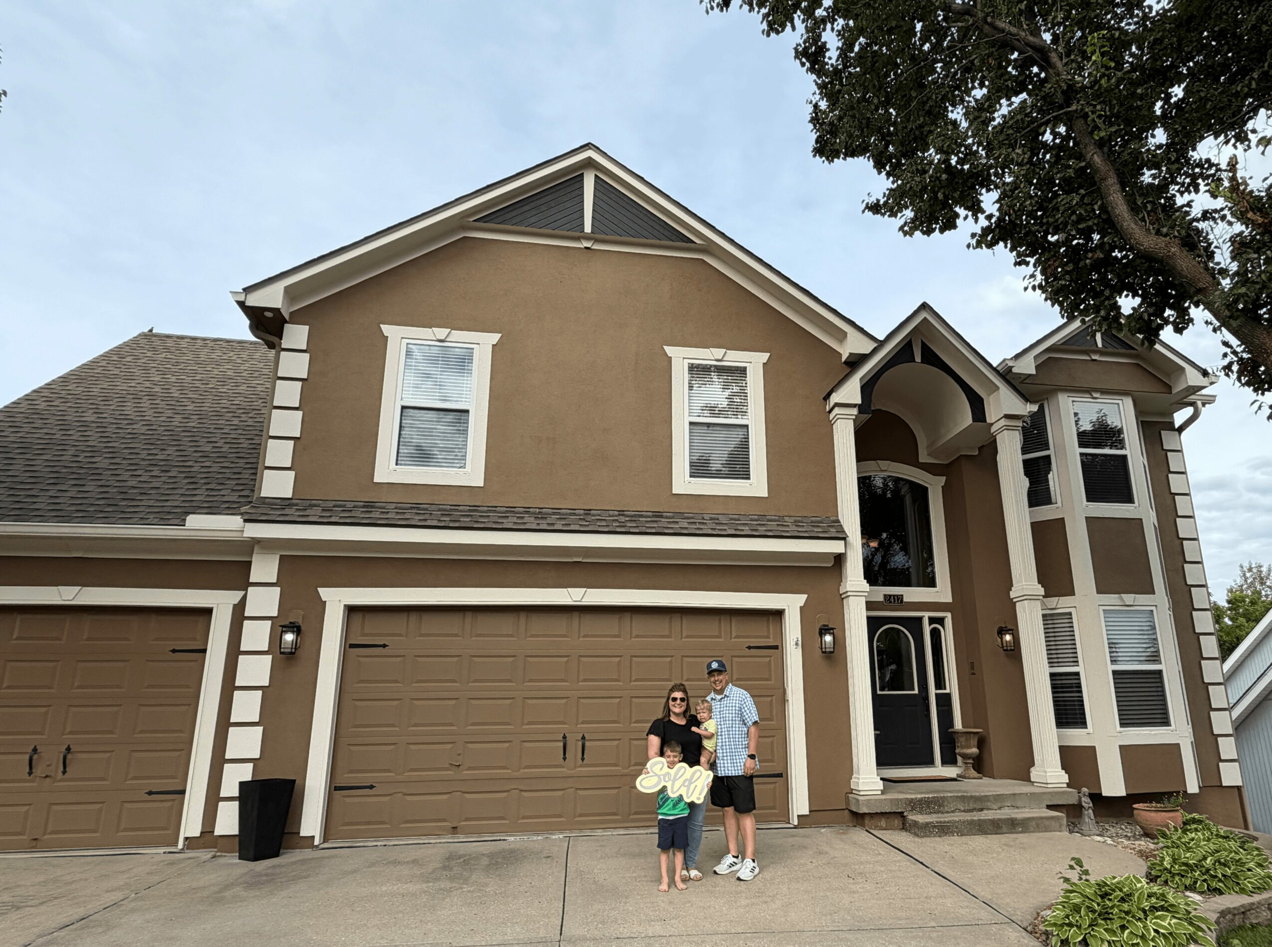 Two children and an adult standing in front of a two-story house with a garage, trees, and a driveway.