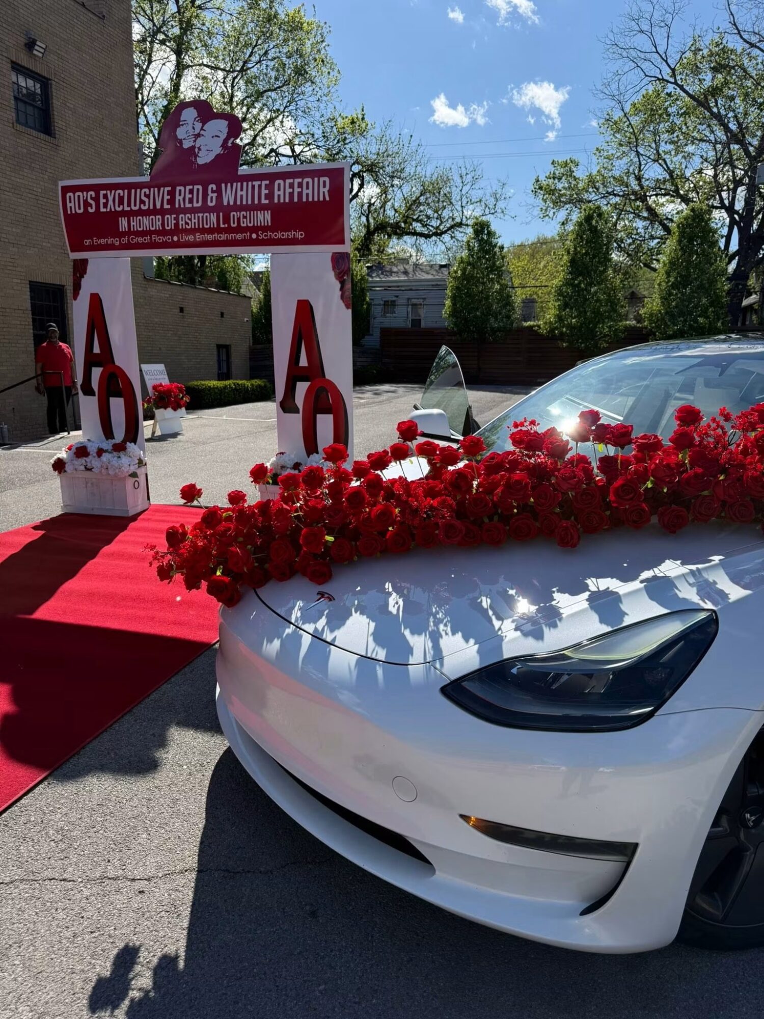 White car decorated with red flowers at a wedding event outdoors, with a red carpet and wedding arch in the background.