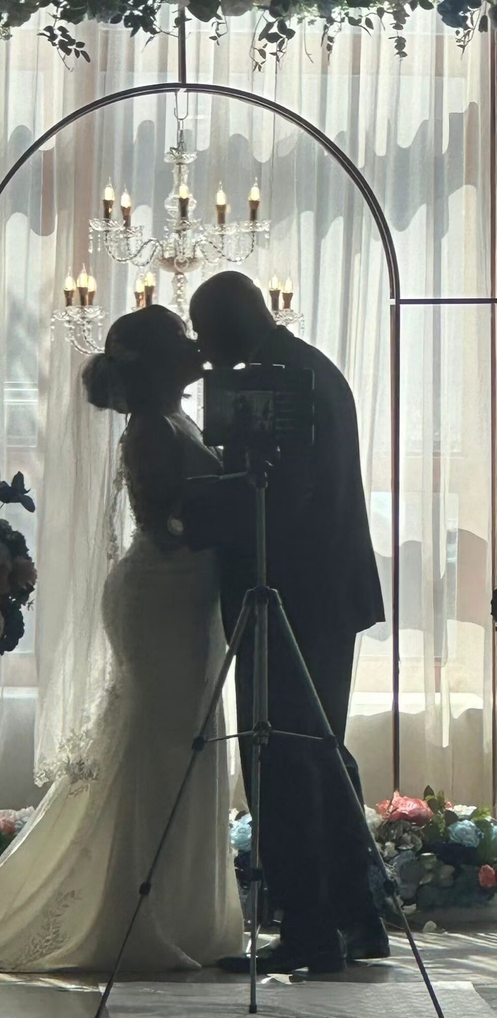 Silhouette of a bride and groom taking a photo in front of a curtain with a chandelier, flowers on the floor.