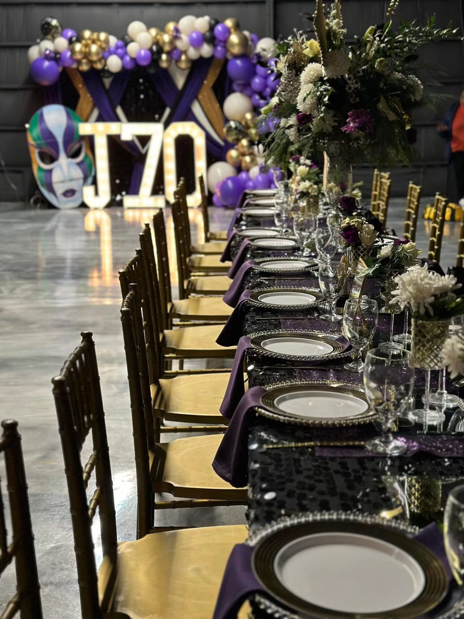 Long table set with plates, glasses, and purple napkins, decorated with floral centerpieces, in a decorated event space with balloons and a skull mask in the background.
