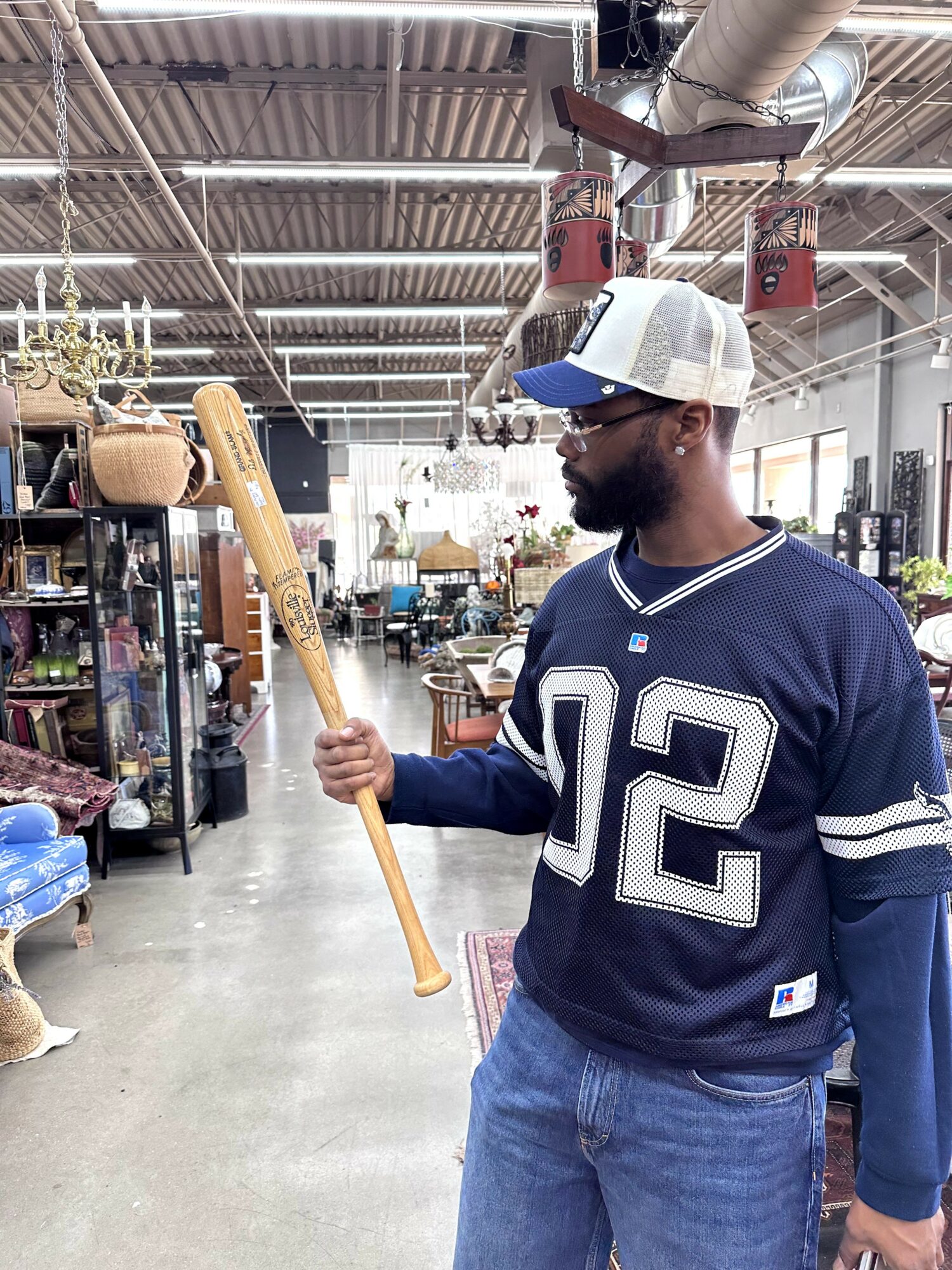 Man in a baseball cap and jersey holding a long wooden stick inside a store with shelves and products.
